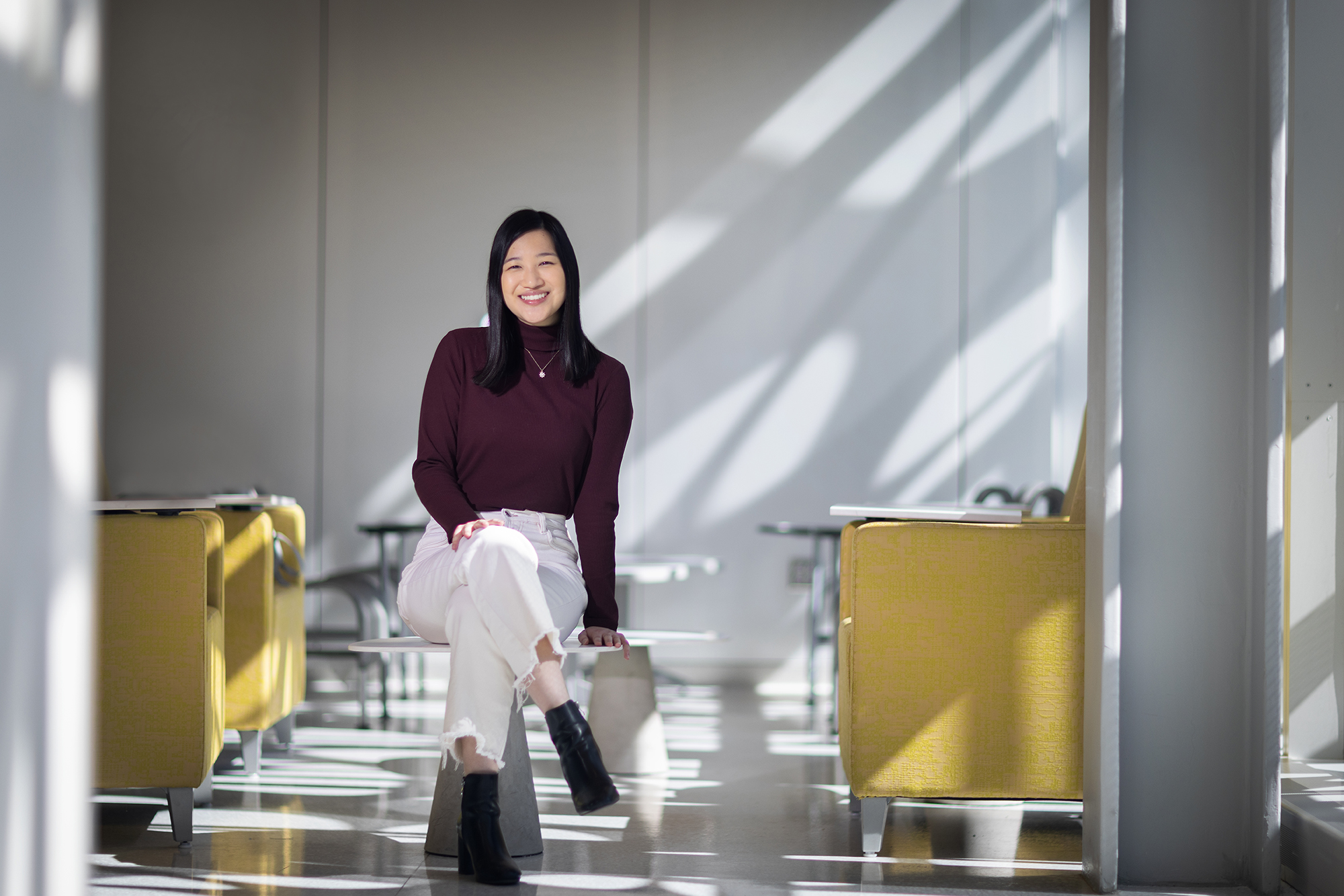 Tiffany Tieu smiles at the camera as she sits on a low white table next to yellow leather chairs.