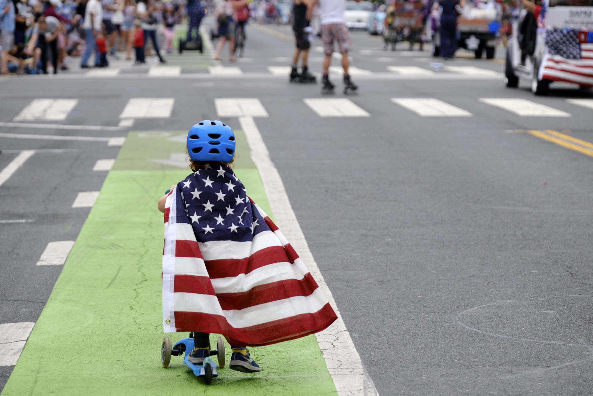A toddler is seen from behind riding a scooter with a blue helmet on and wearing an American flag like a cape.