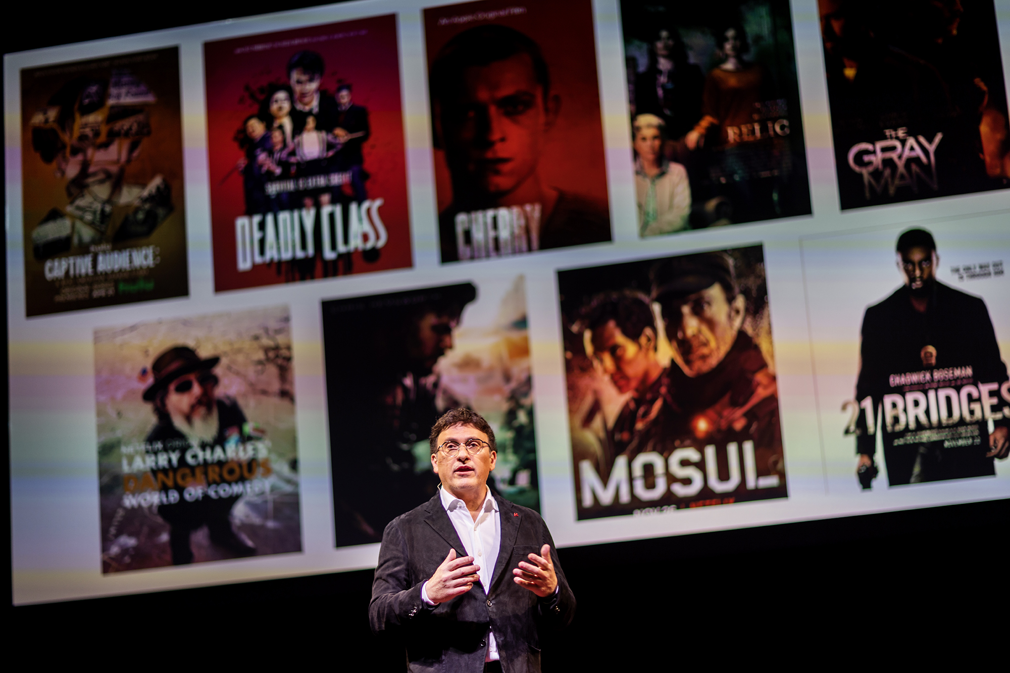 Anthony Russo speaks on stage with movies he's directed in the background