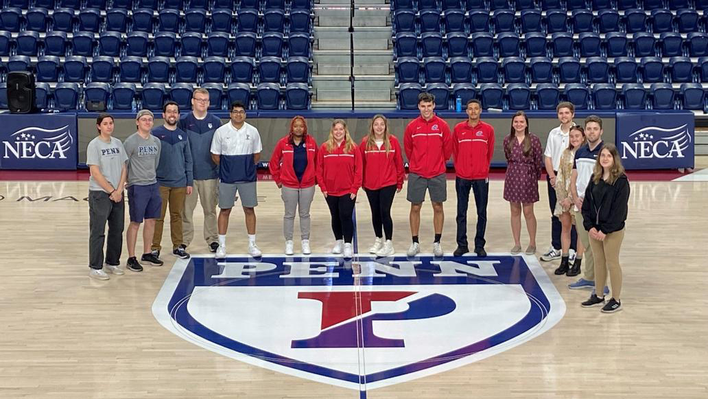 A group of Penn Athletics interns stand at center court at the Palestra near the Penn logo.