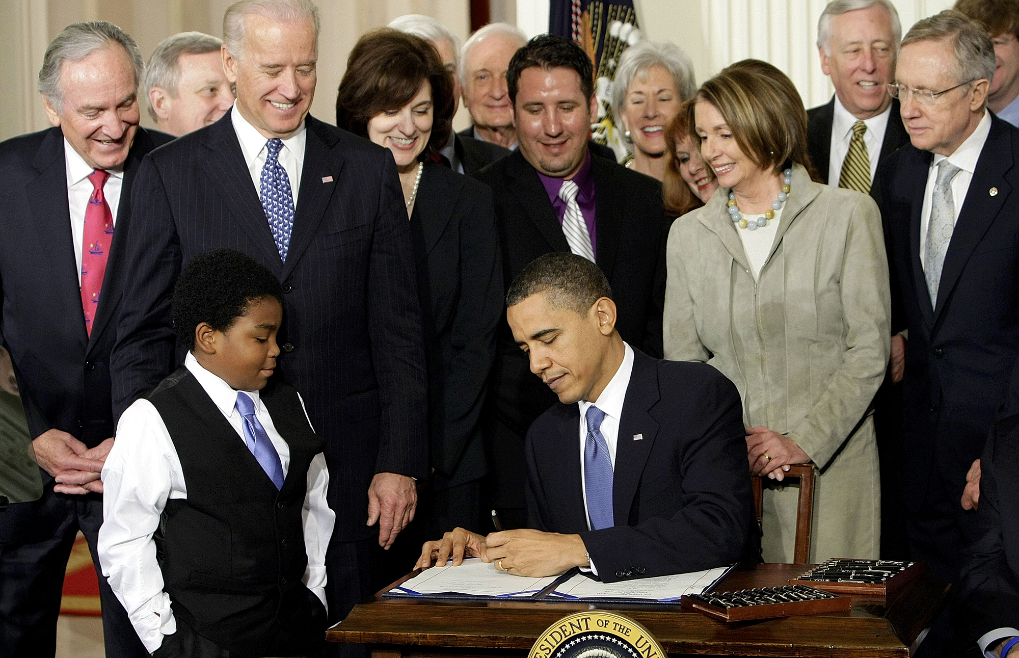 President Barack Obama signs the Affordable Care Act surrounded by lawmakers and a young child standing by the table.