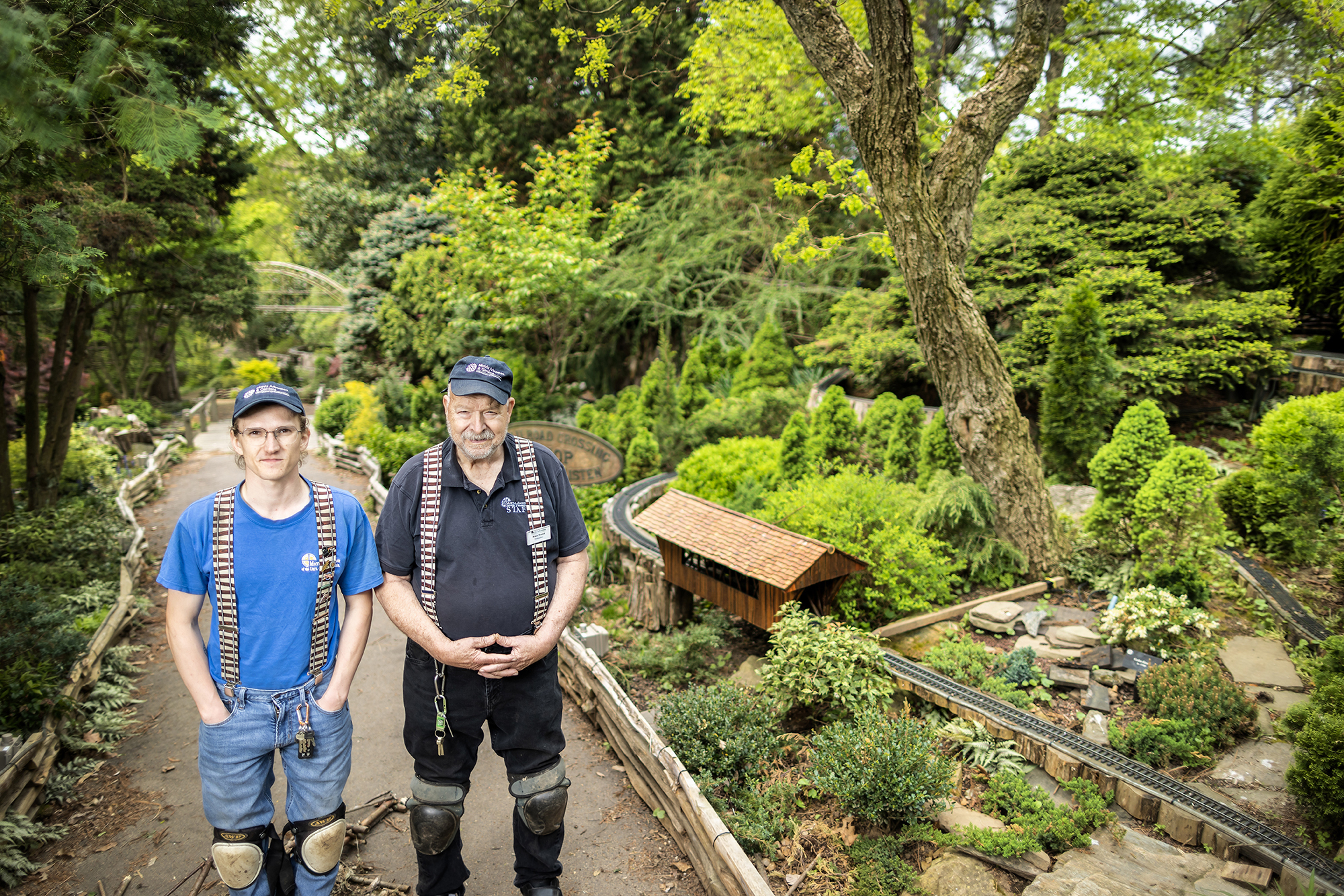 Josh Faia and Bruce Morrell standing next to garden railway surrounded by trees and foliage. 