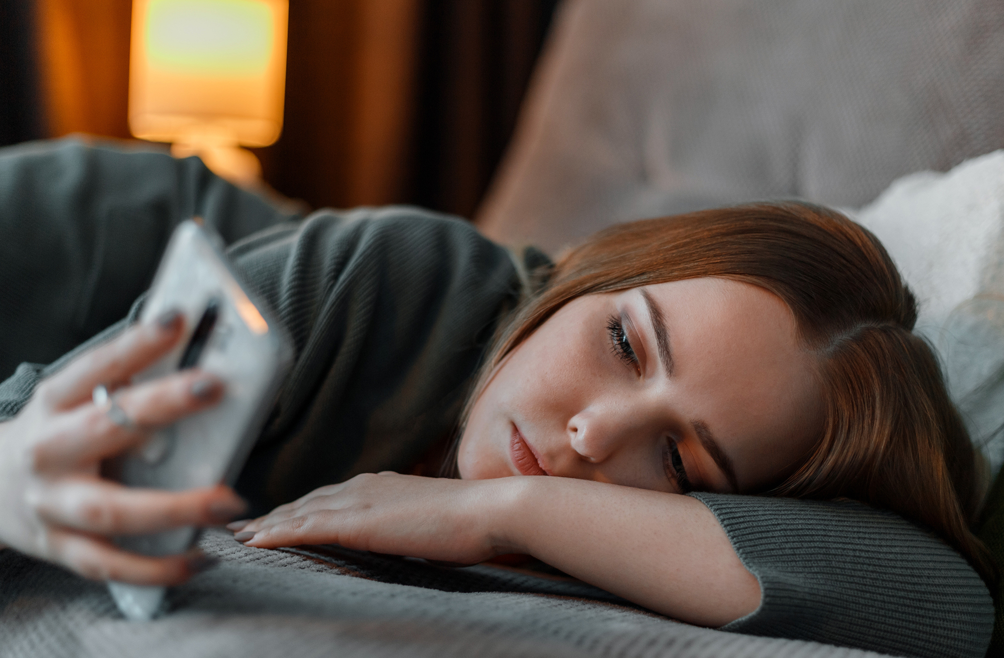 A young woman lies sideways on a bed, gazing at her phone