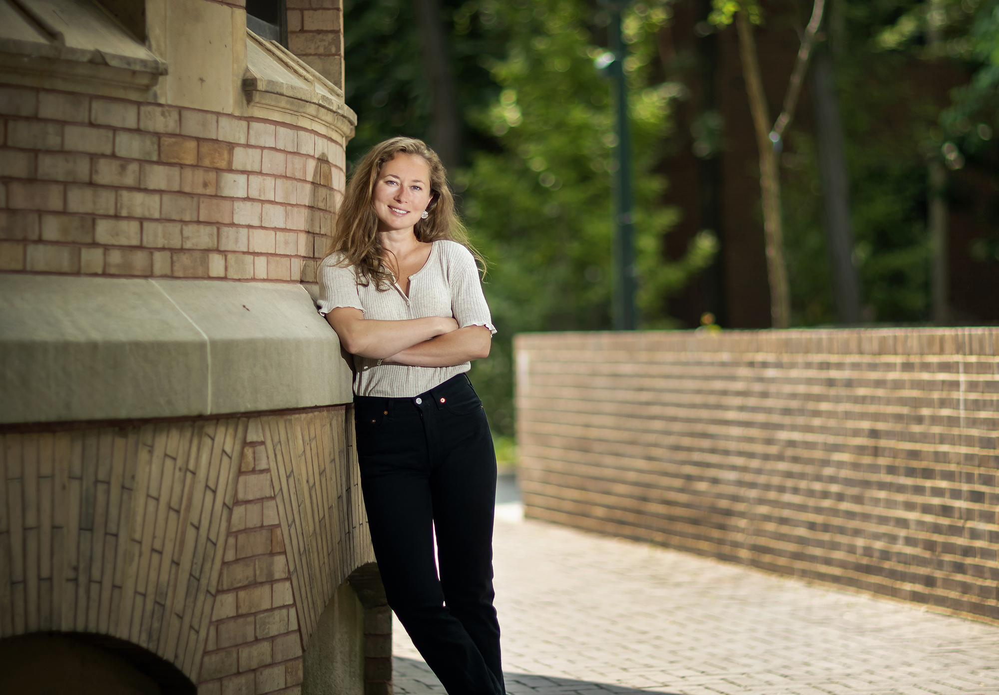 History Ph.D. candidate Arielle Alterwaite leans up against a light brown brick building with her arms crossed, smiling at the camera.