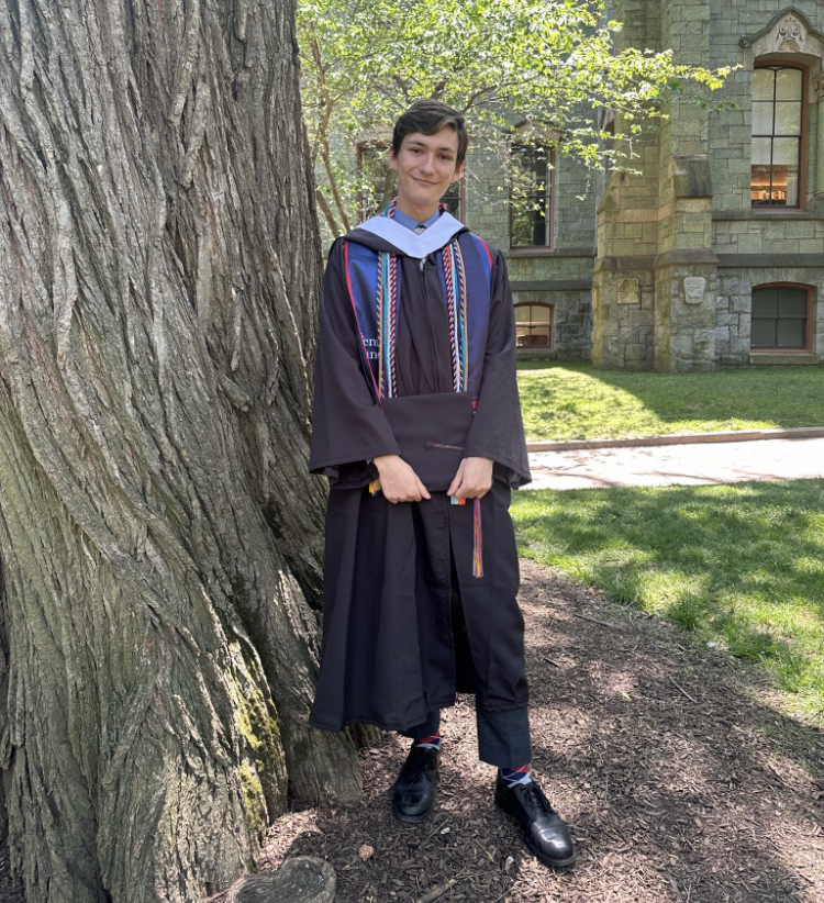 Justin Roberts in cap and gown, standing against a tree