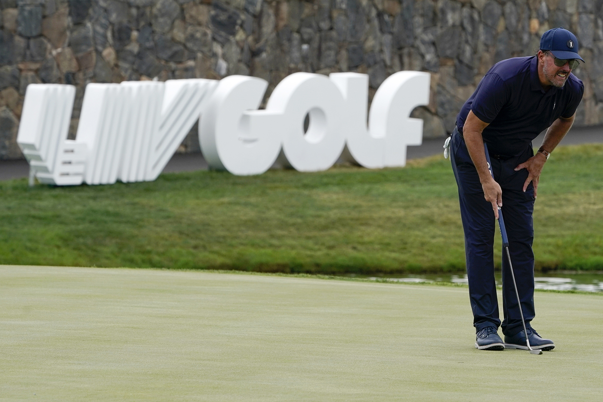 Golfer Phil Mickelson holds a golf club standing on grass and looks into the distance, bending at the waist, in front of a LIV Golf sign and a rock wall.