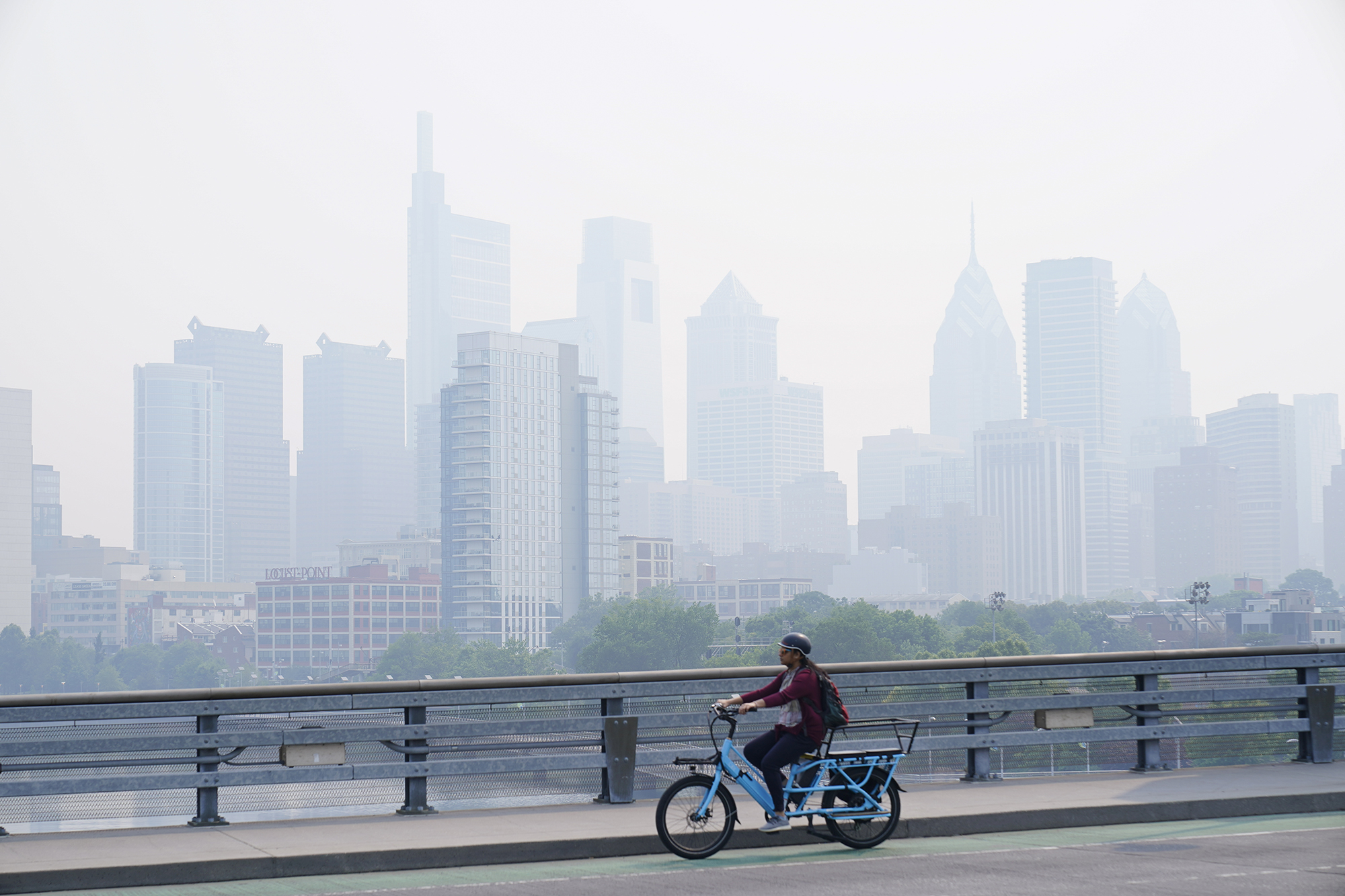 A person crosses the Schuylkill River on a blue bike. The city skyline behind him is obscured with smoke haze.