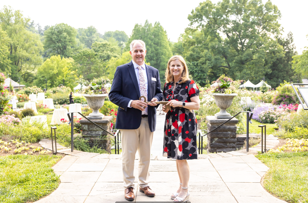 Bill Cullina and Liz Magill stand holding a large ornamental key in the Morris' rose garden.