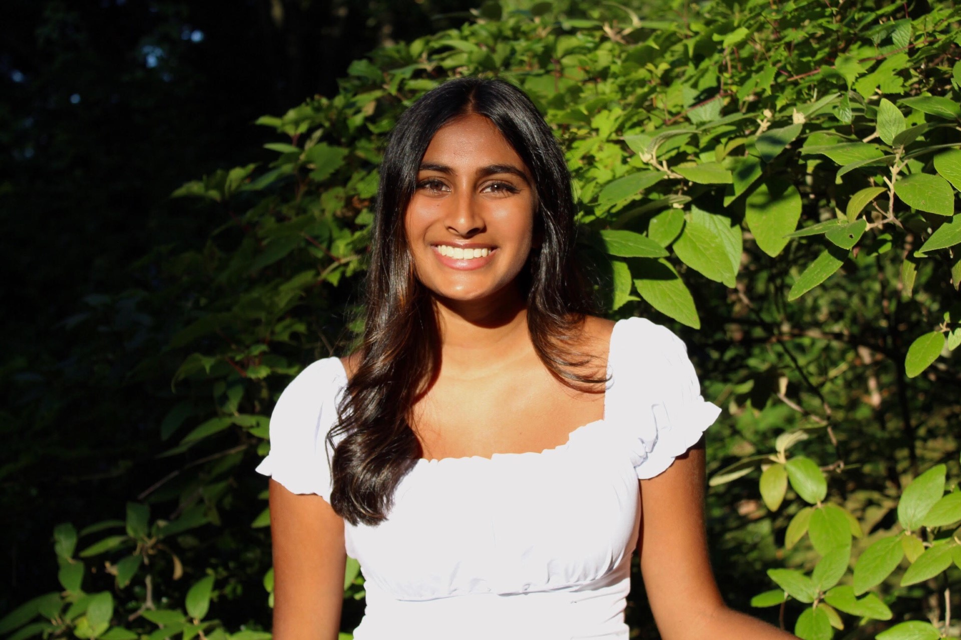 Ayesha Patel standing in outside in front of a leafy bush.