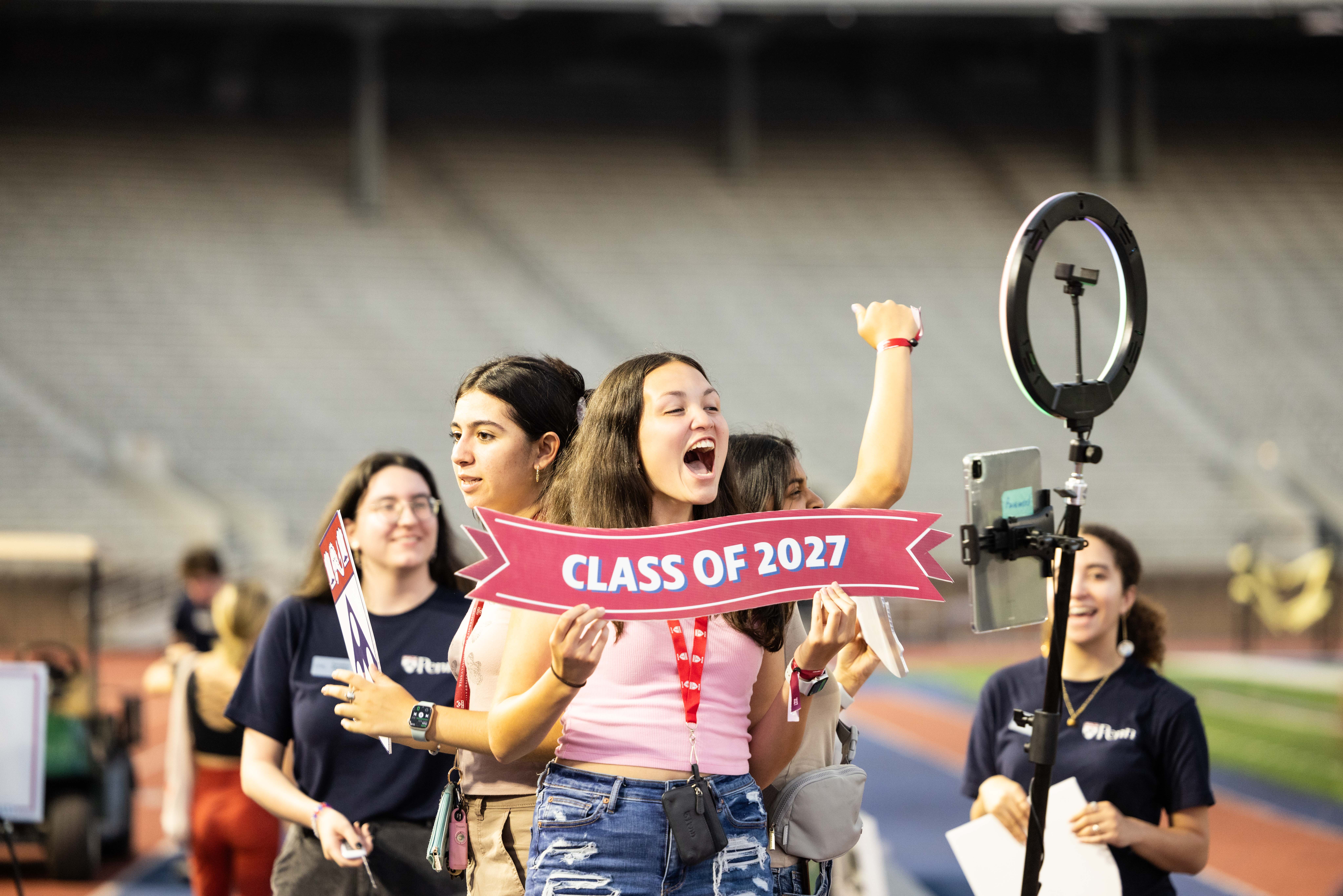 A woman holding a sign reading "Class of 2027" cheers while speaking into a camera