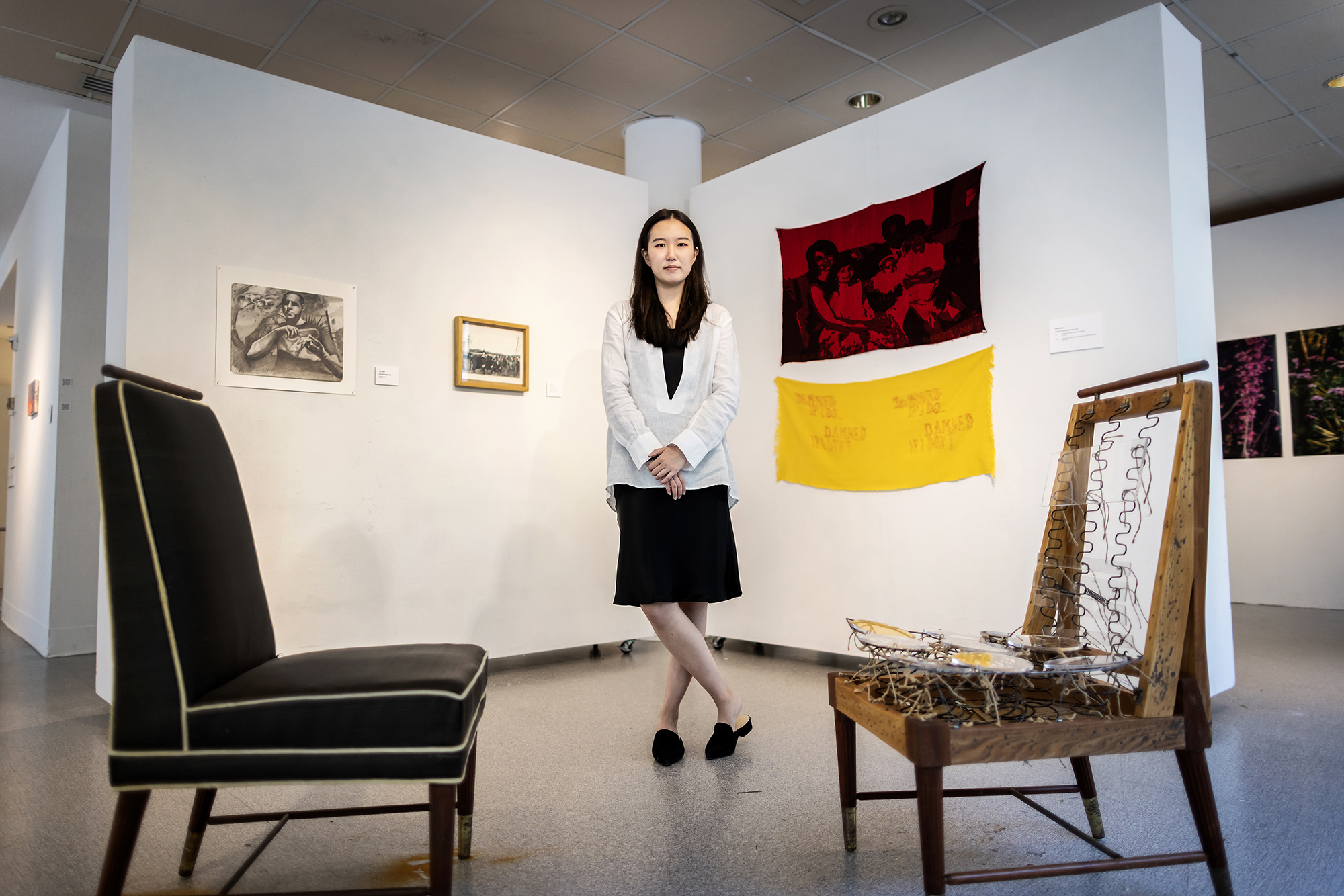 Kay Seohyung Lee standing in art gallery with an installation with two chairs in front of her and a wall with four artworks behind her.