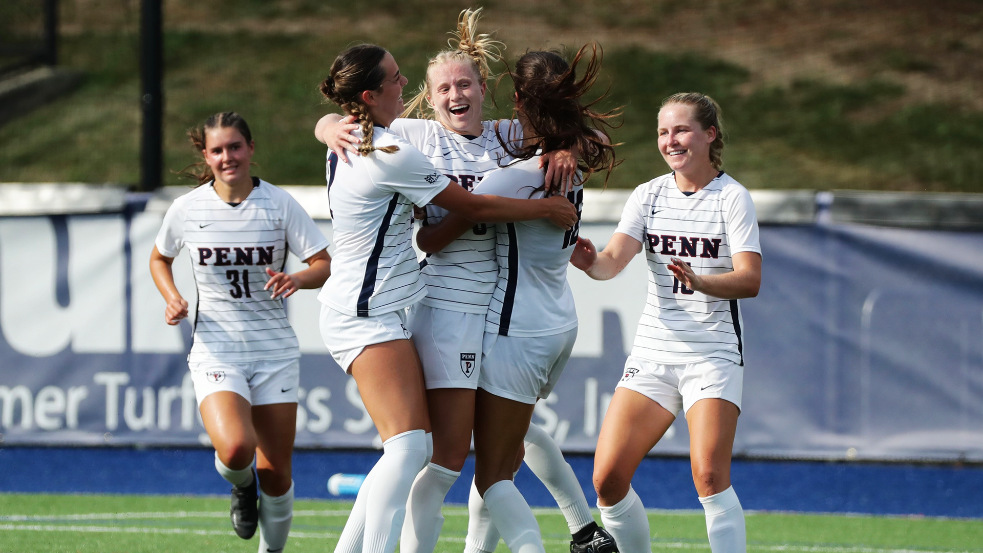 Women's soccer players hug and celebrate after a big moment during a game.
