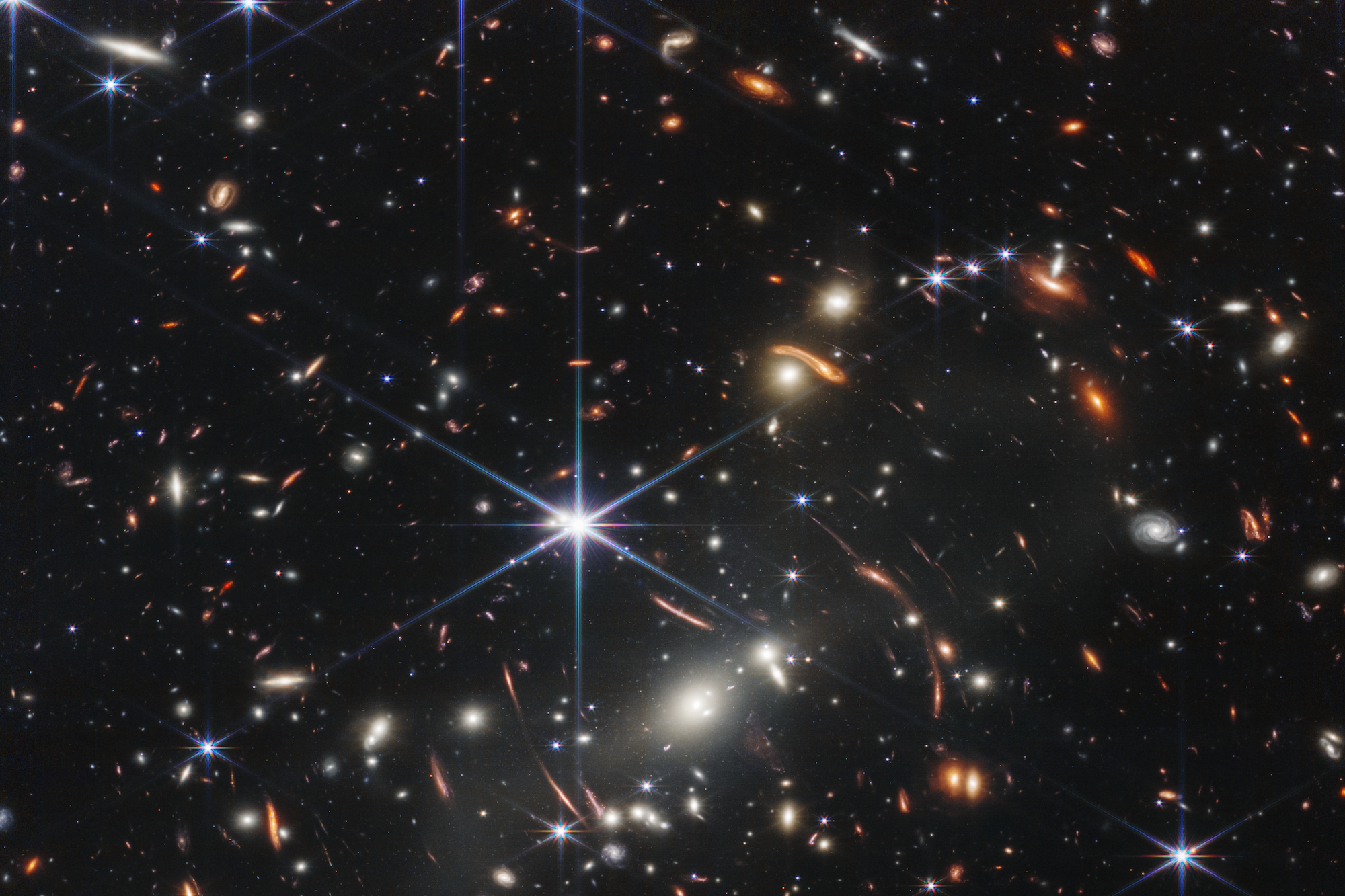 Thousands of small galaxies appear across this view. Their colors vary. Some are shades of orange, while others are white. Most appear as fuzzy ovals, but a few have distinct spiral arms. In front of the galaxies are several foreground stars. Most appear blue, and the bright stars have diffraction spikes, forming an eight-pointed star shape. There are also many thin, long, orange arcs that curve around the center of the image.