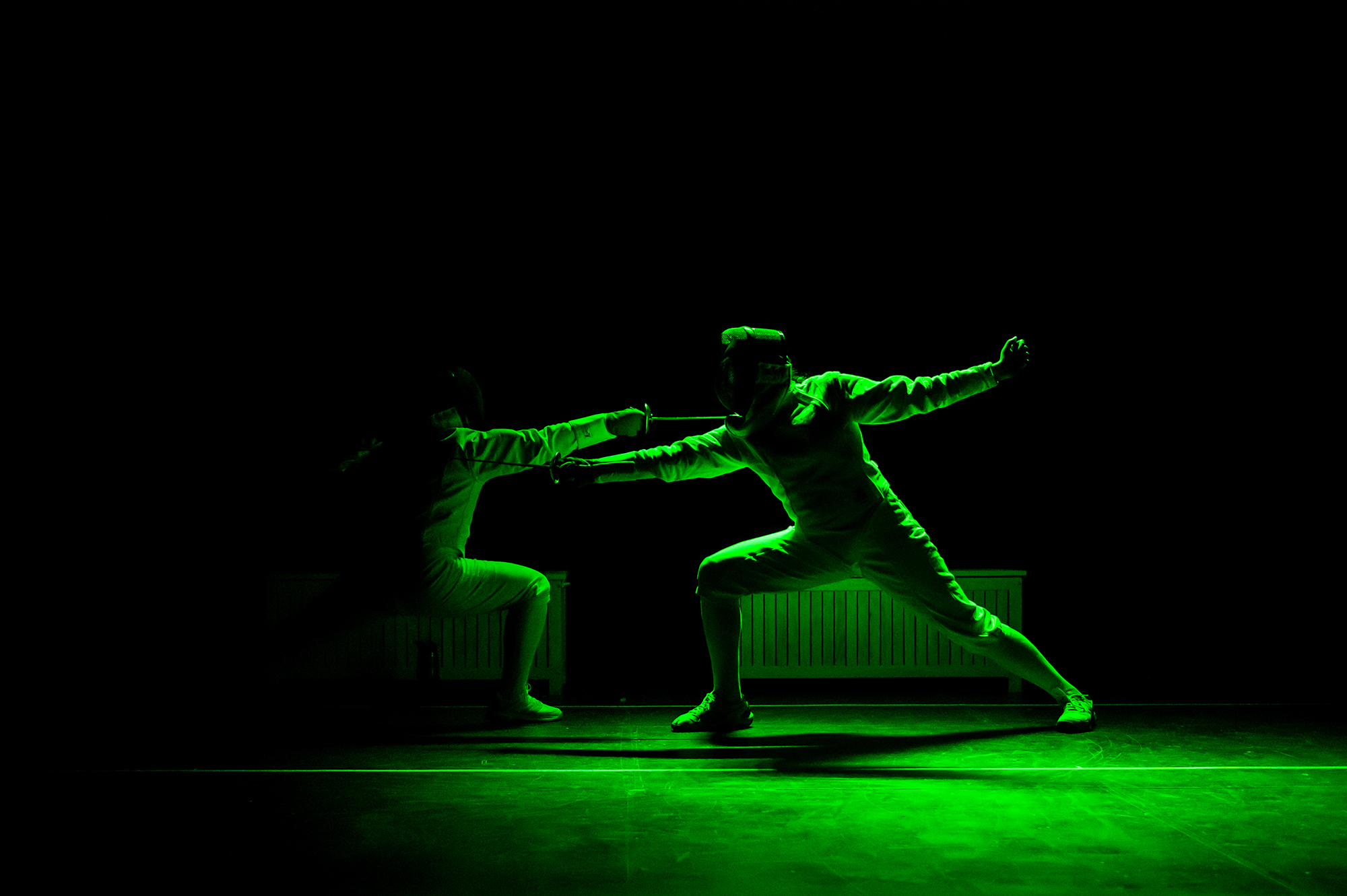 Two actors in fencing gear performing on stage in the dark with green light.