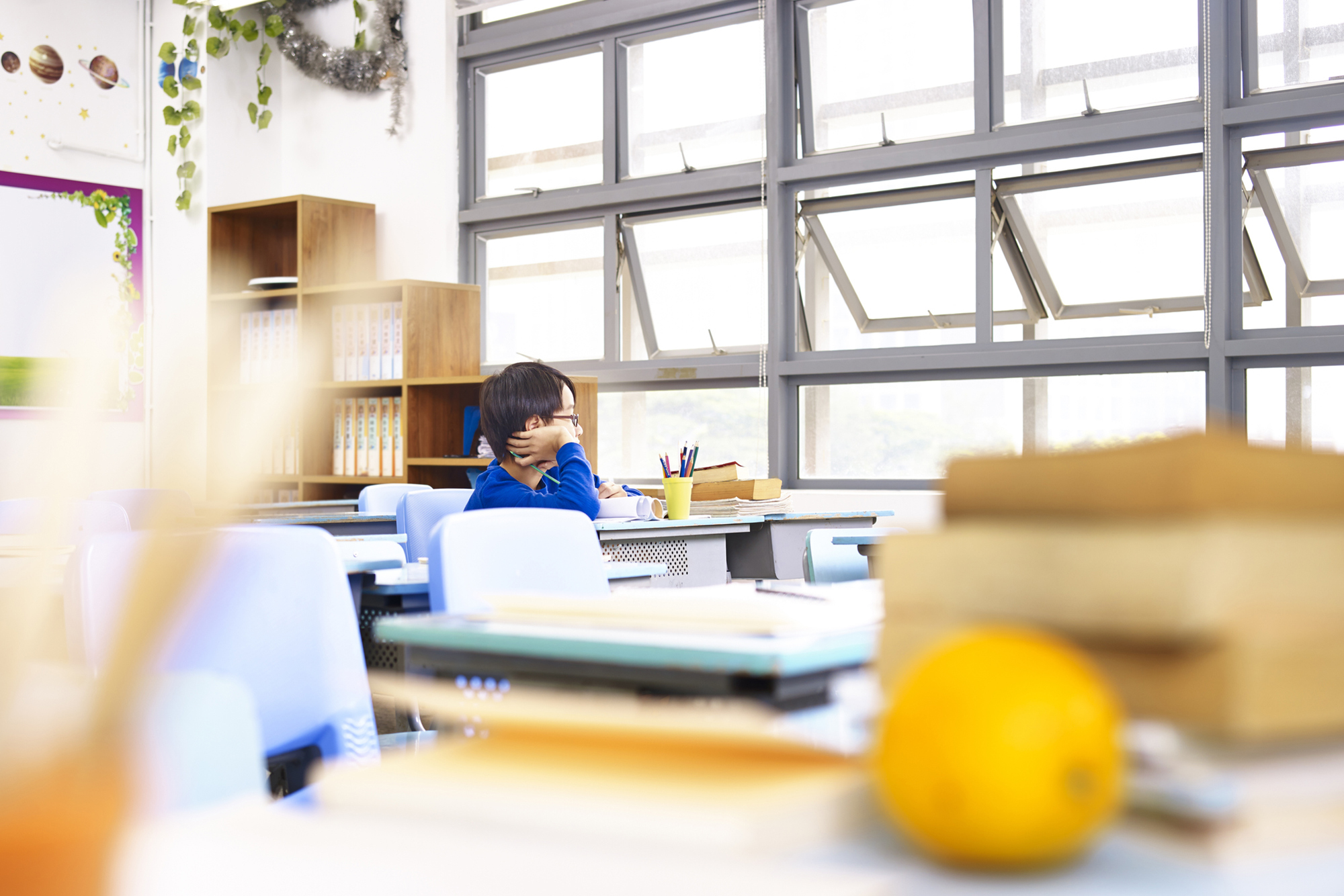 A young student looks out the window of a classroom with several empty desks.