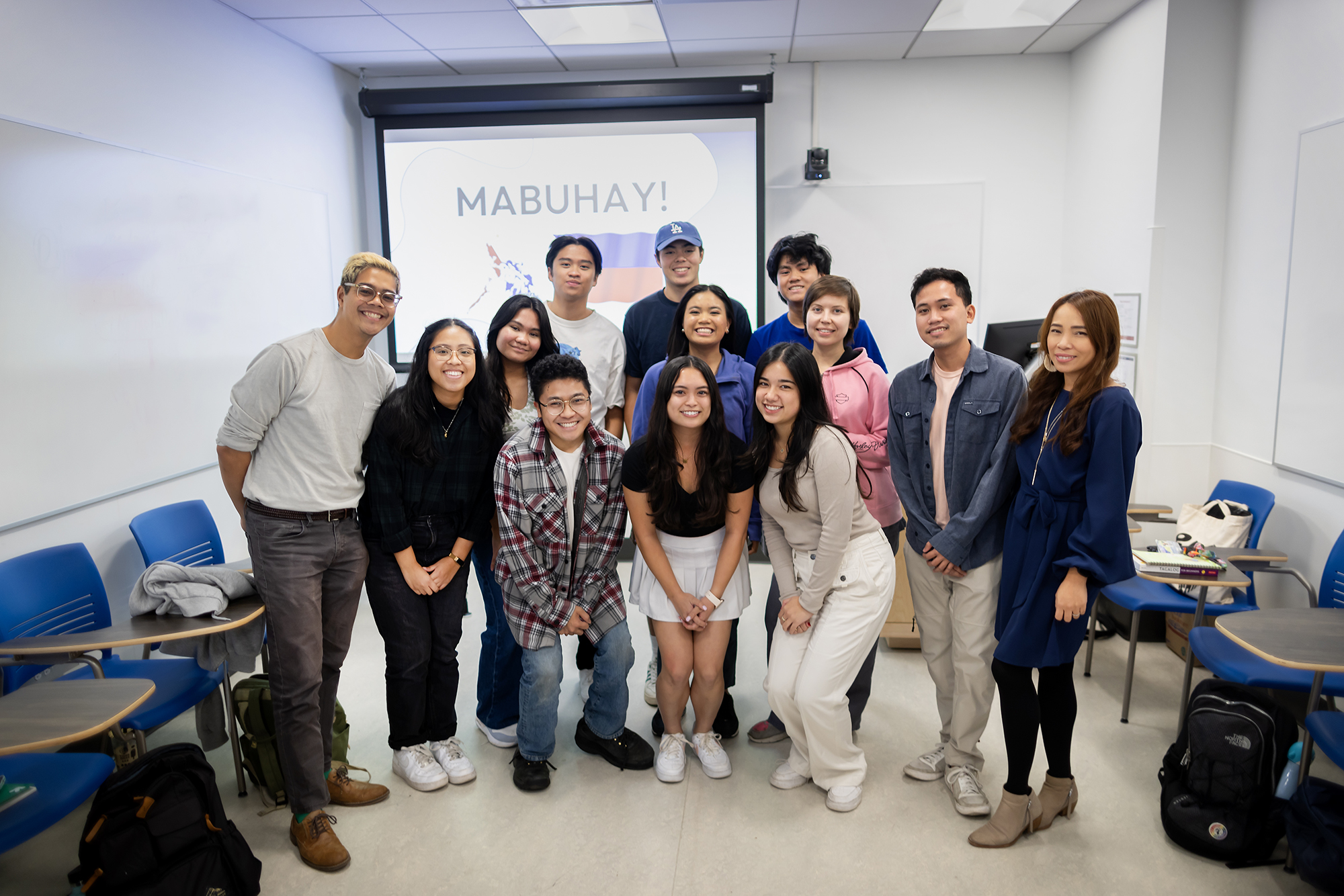 A group of students stand with their professor and teaching assistance. A word on the screen behind them reads, "Mabuhay!" meaning long life.