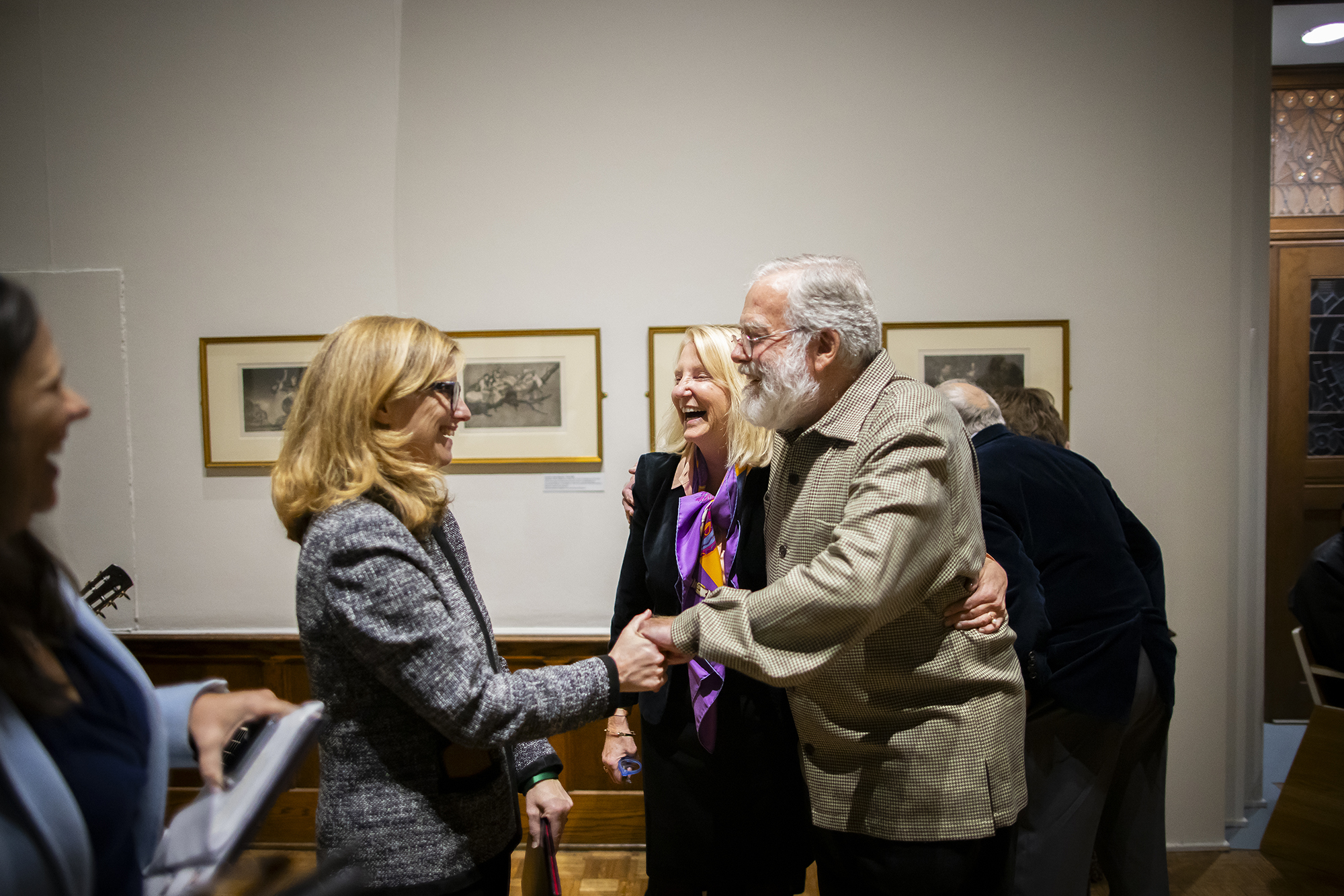Two people shaking hands with another laughing in background at an art gallery exhibition