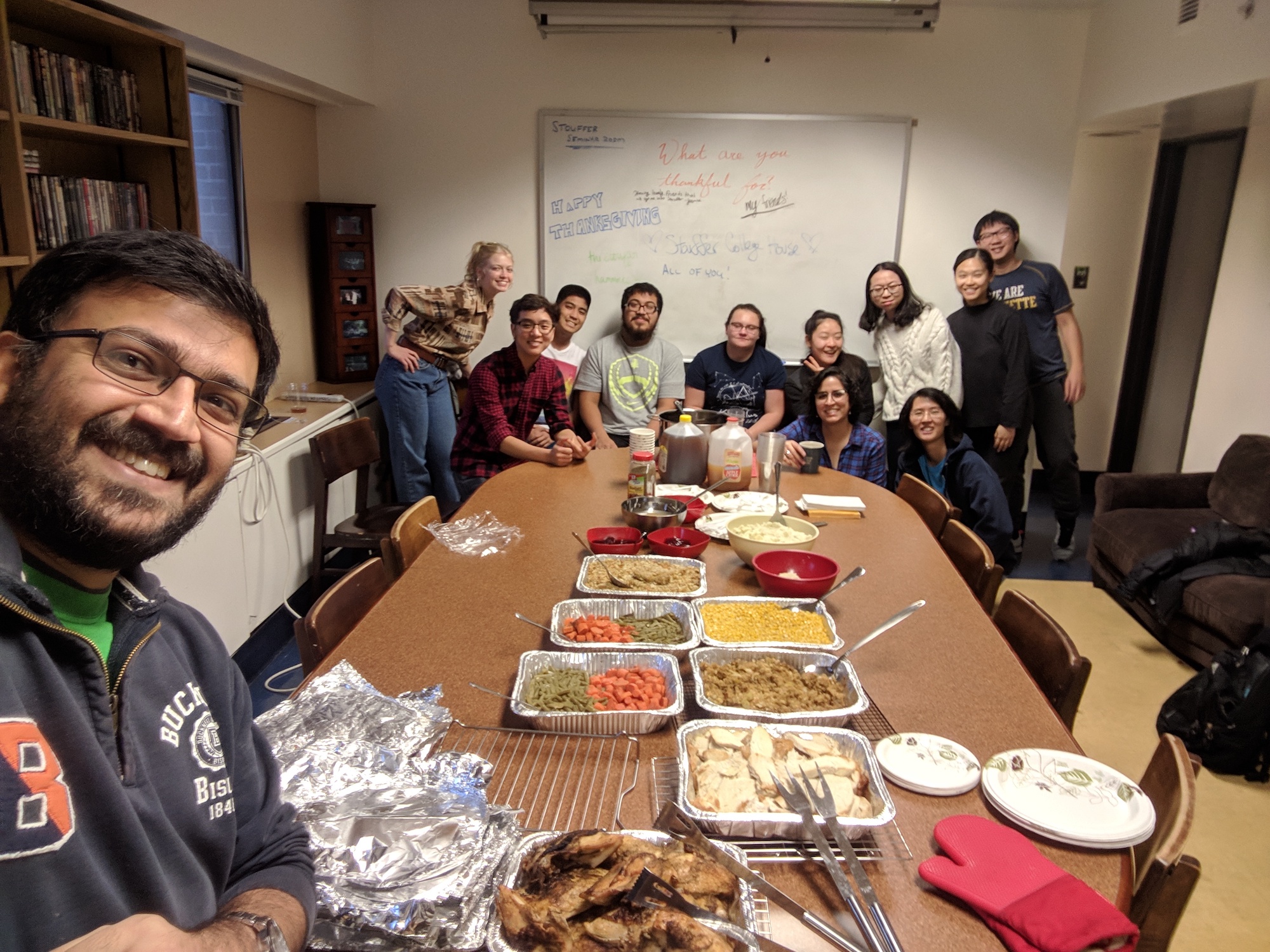 Nadir Sharif takes a selfie with a table full of Thanksgiving food and a group of college students behind him. A whiteboard at the back of the room reads "What are you thankful for?"