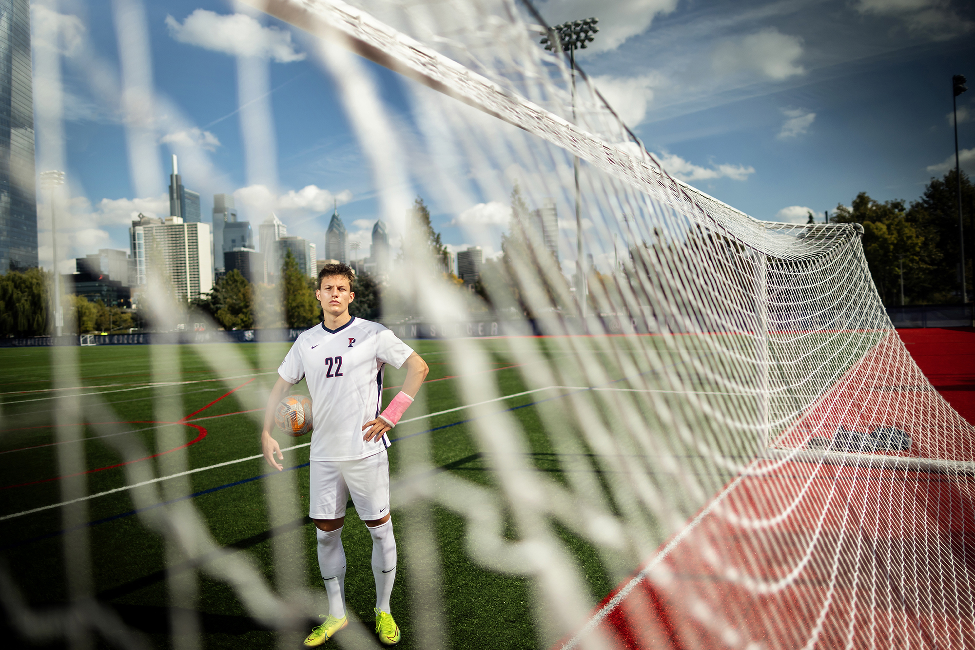 Stas Korzeniowski stands on the field at Penn Park with a soccer ball on his right side, with the Philly skyline in the background.