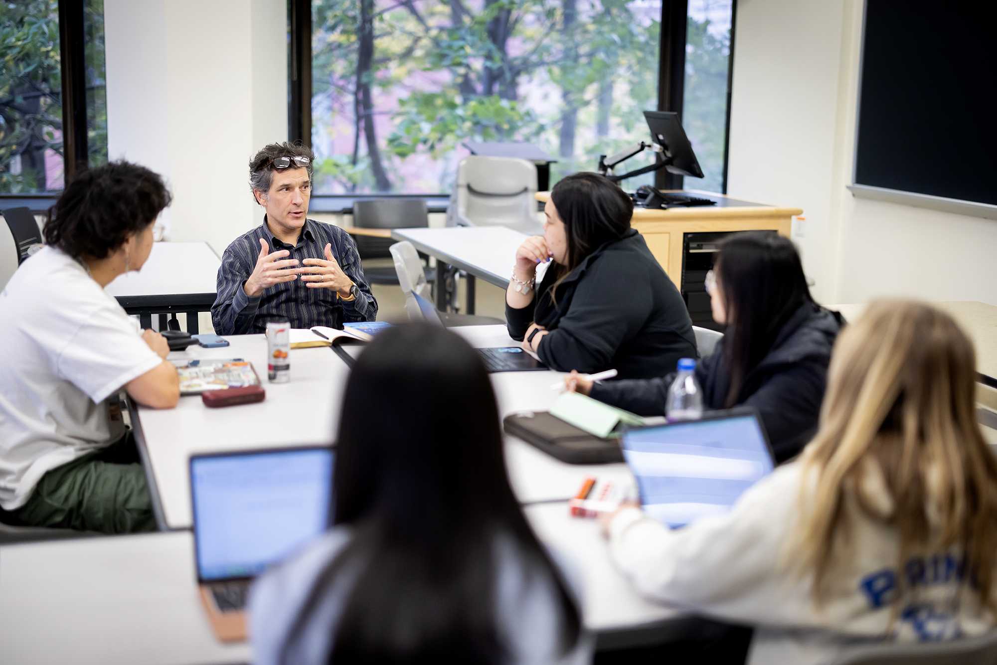 Tyson Smith gestures at the head of a table full of students, in front of windows showing trees.