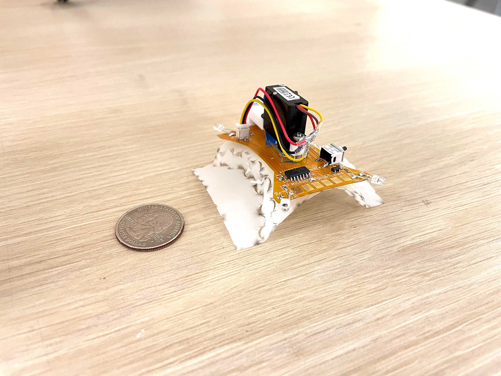A centimeter-scale quadruped robot sitting on a desk next to a a quarter for scale