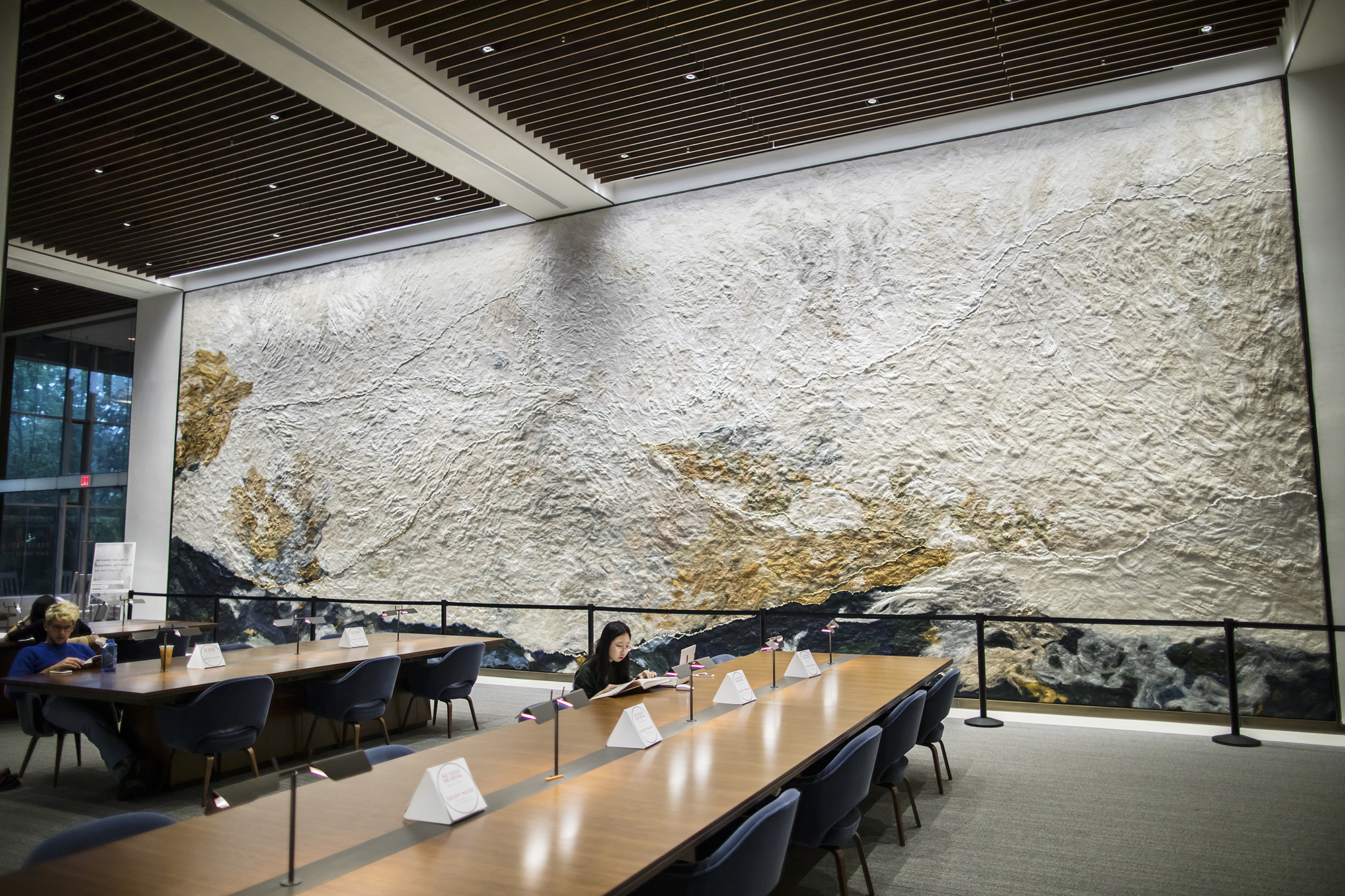 An image of the Moelis Reading Room, where students study at long tables. In the background, a textile mural is shown. 