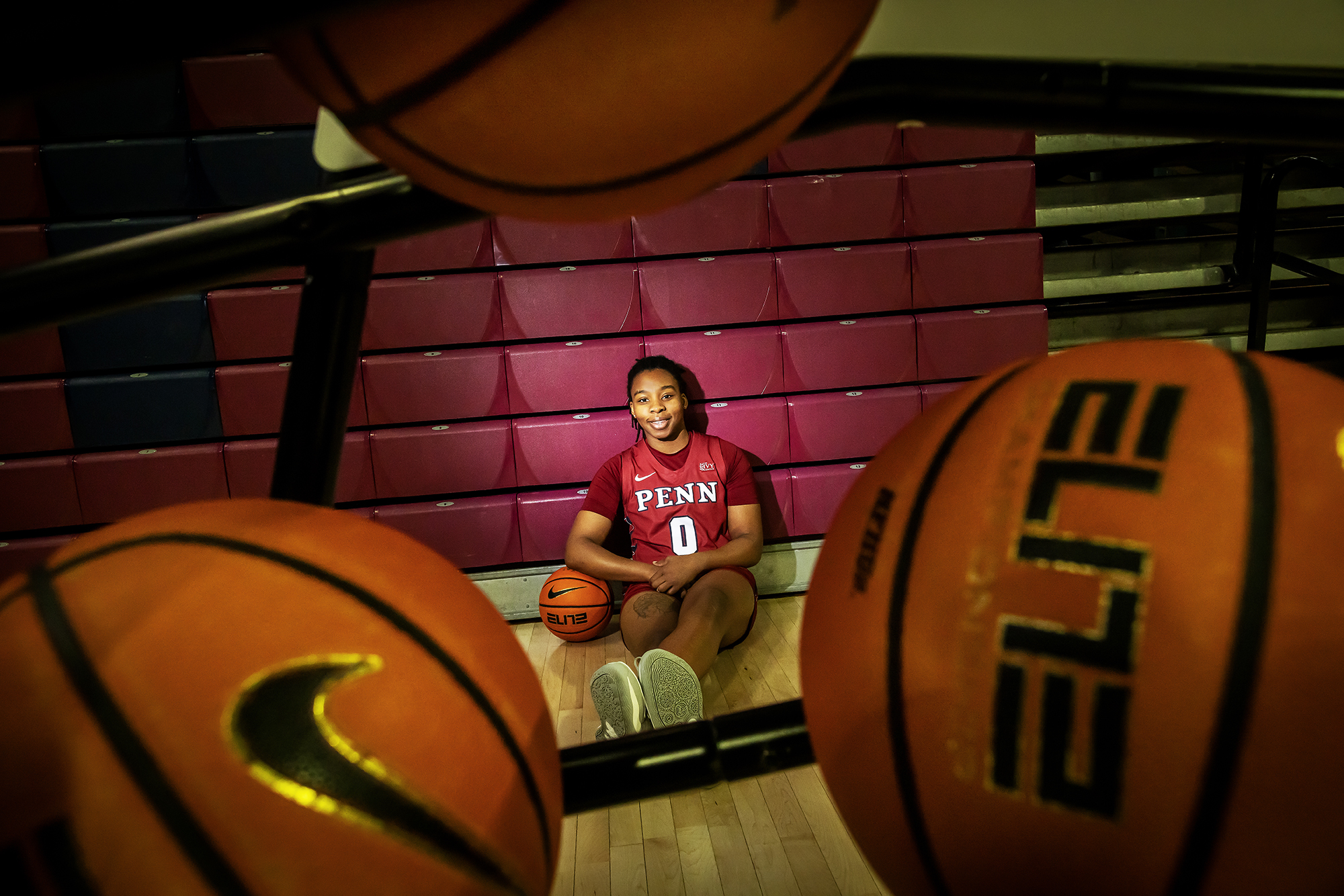 Jordan Obi sits on the court with her elbow rested on a basketball, wearing her red Penn jersey.