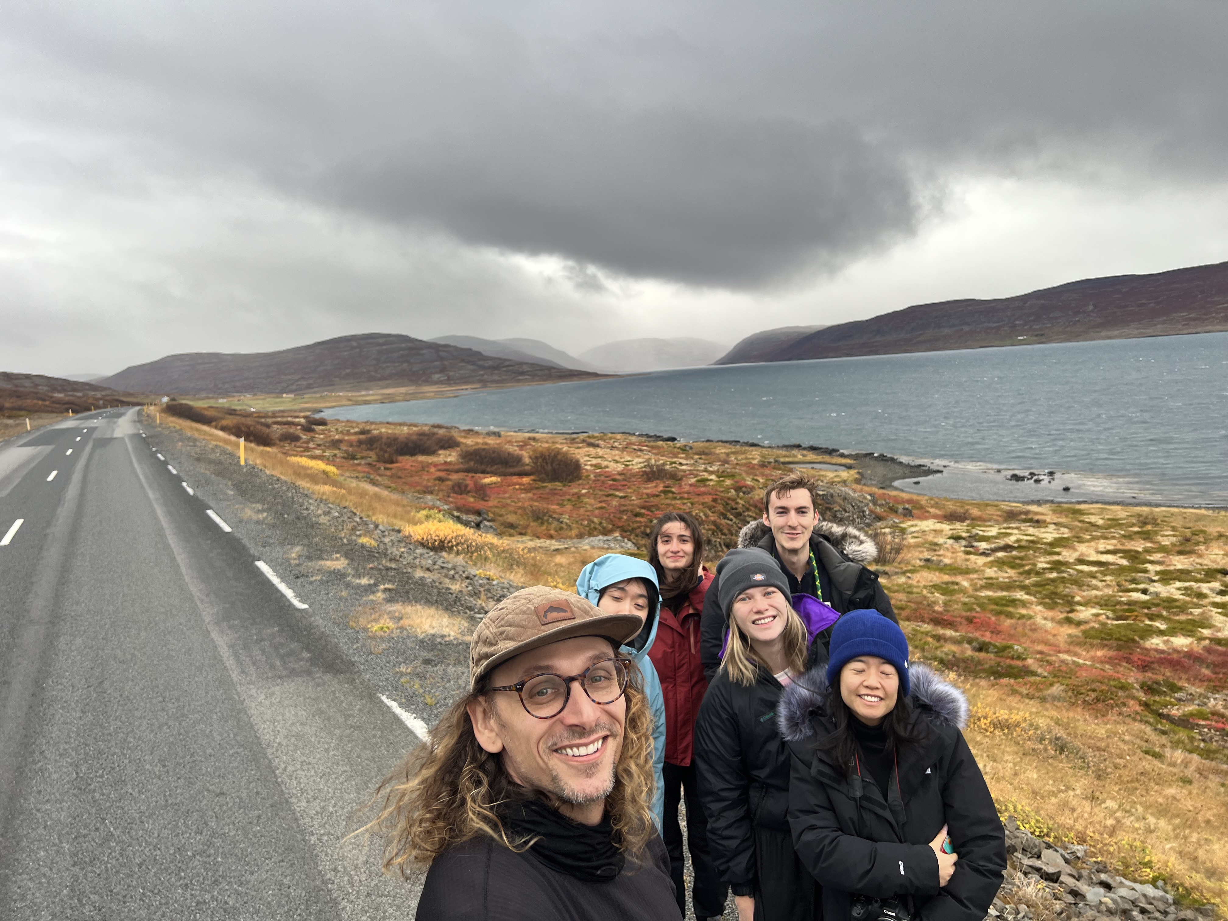 Six people take a group selfie by the side of the road in Iceland, with mountains and water in the distance