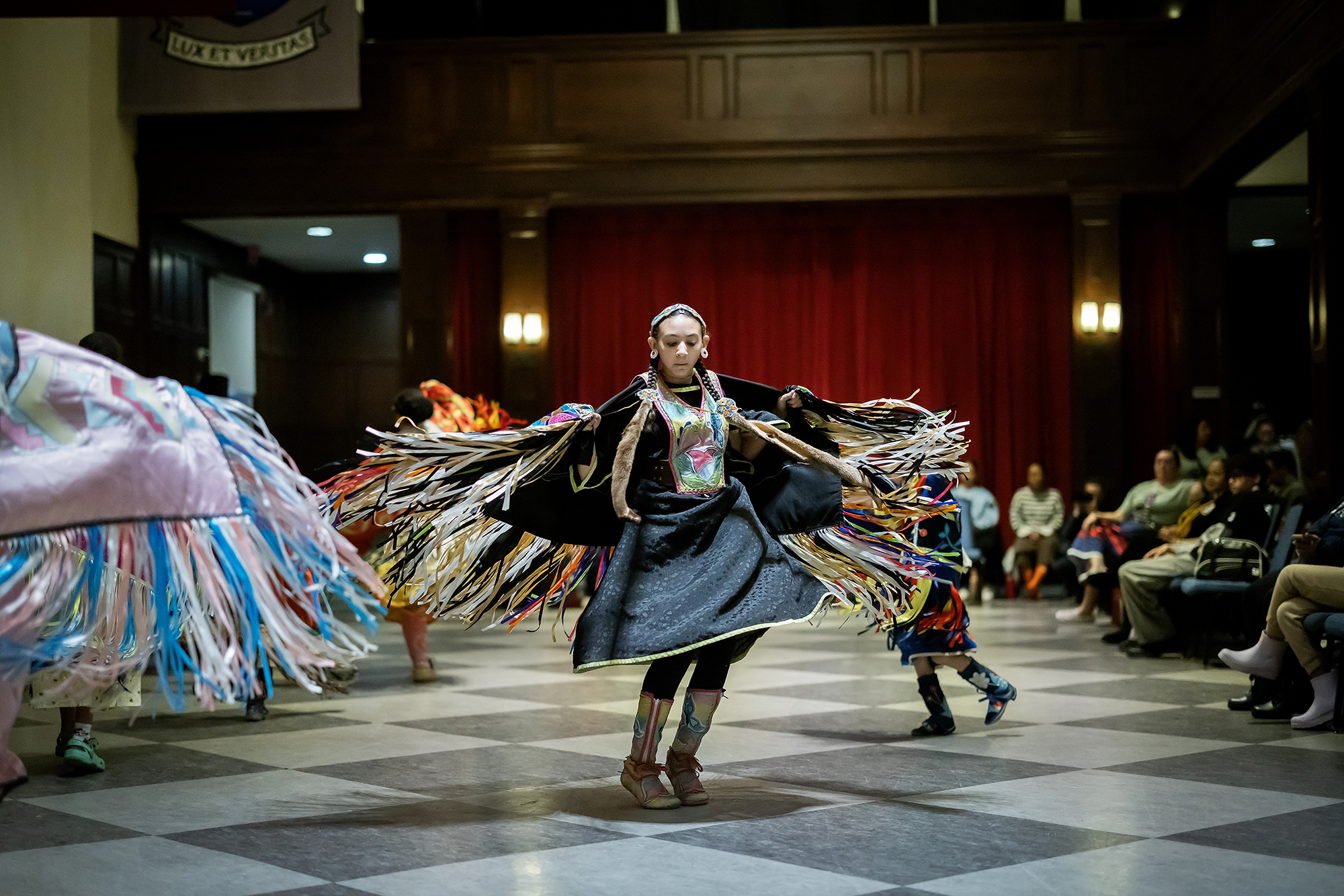 A young woman dances in the center of the hall of flags, with swirling fringed shawls around her.