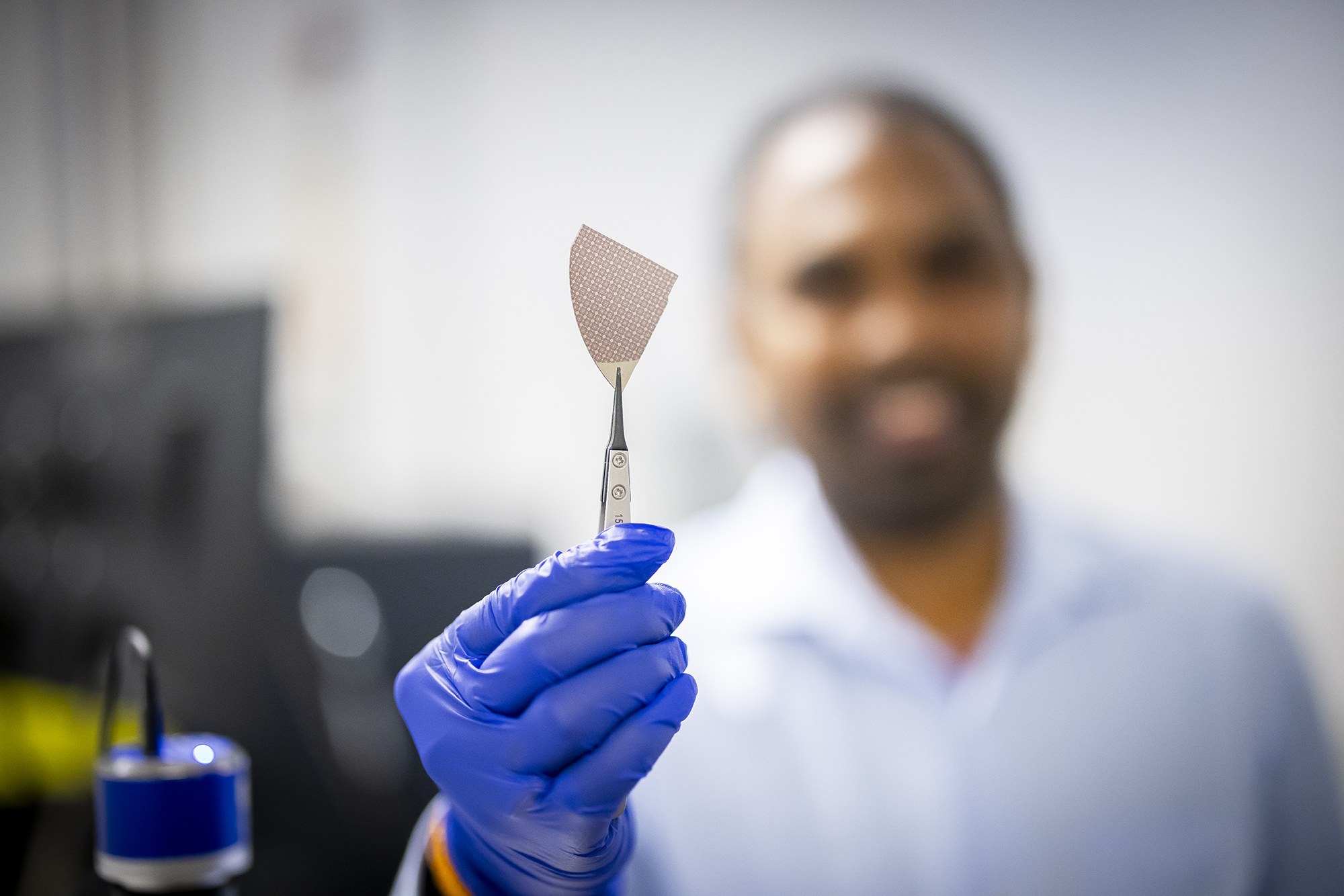 Researcher, out of focus and in the background, holding a pinching tool clasping a memory storage device.