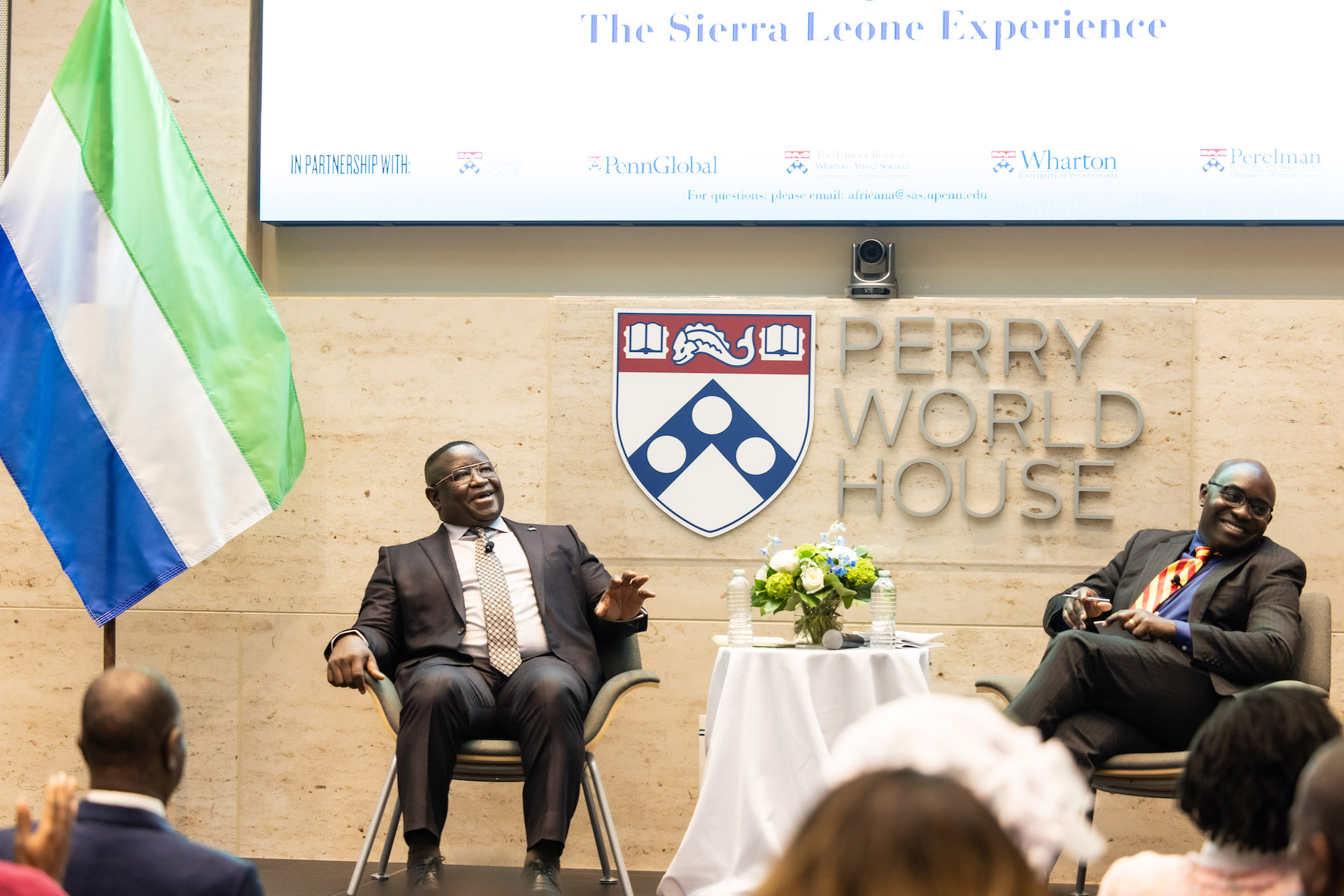 The President of Sierra Leone sits on a stage next to a flag of his nation, and the head of Penn's Center for Africana Studies sits near him and laughs at what he's saying.