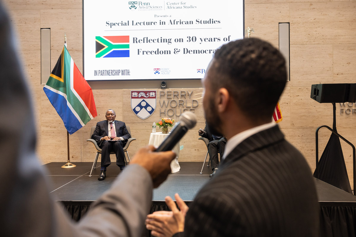 A person holds a microphone up for an audience member who is asking a question of the former president of South Africa, who sits on a stage next to his nation's flag.