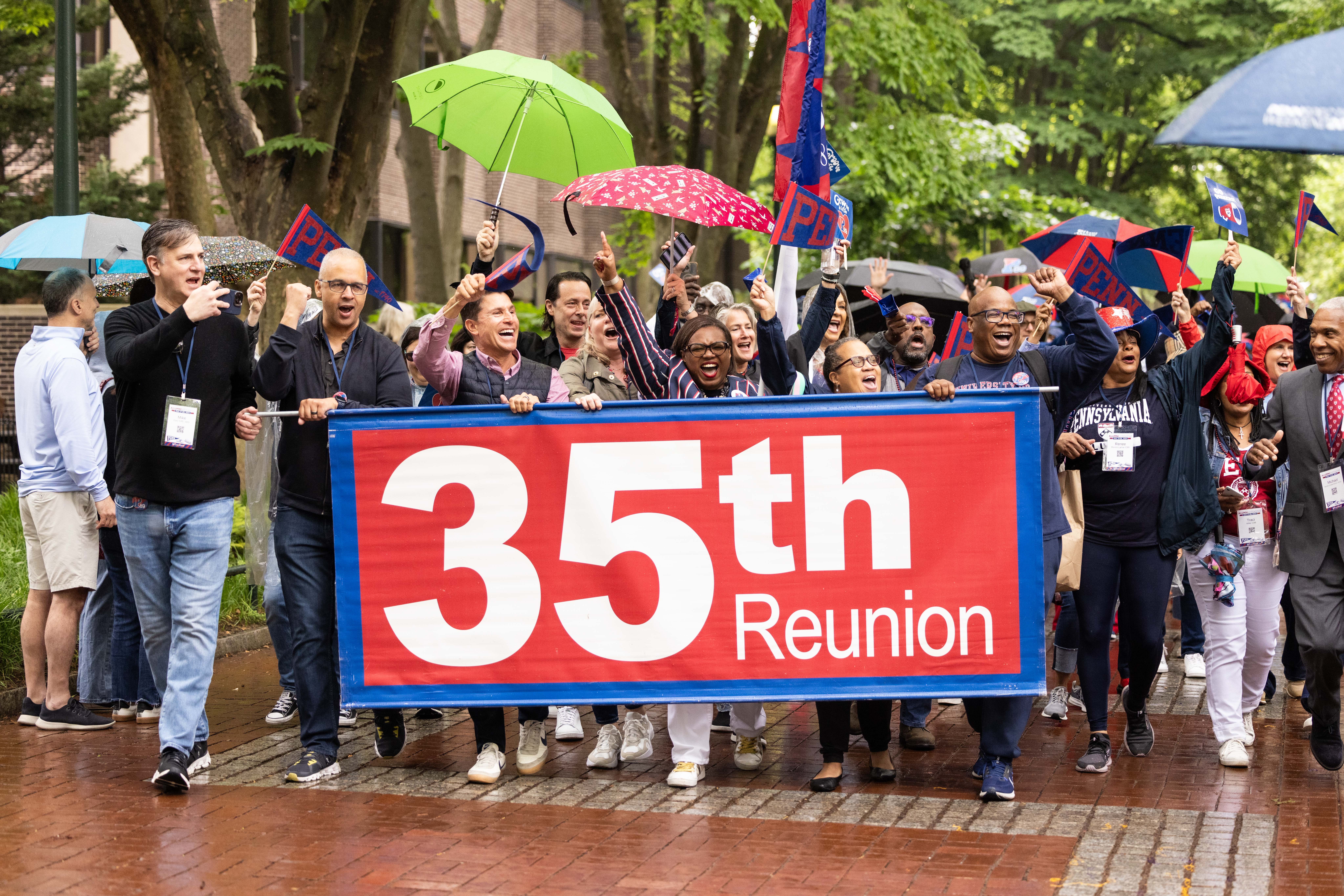 The Class of 1989 march together on Locust Walk during the Alumni Parade, holding a 35th reunion sign and umbrellas