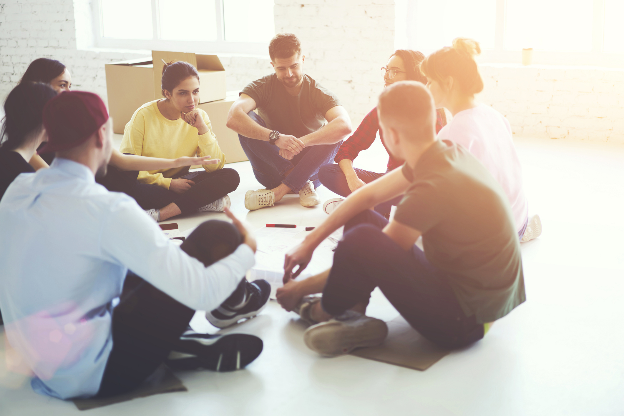 A group of young adults sitting in a circle on the floor, engaged in a collaborative discussion in a bright, spacious room.