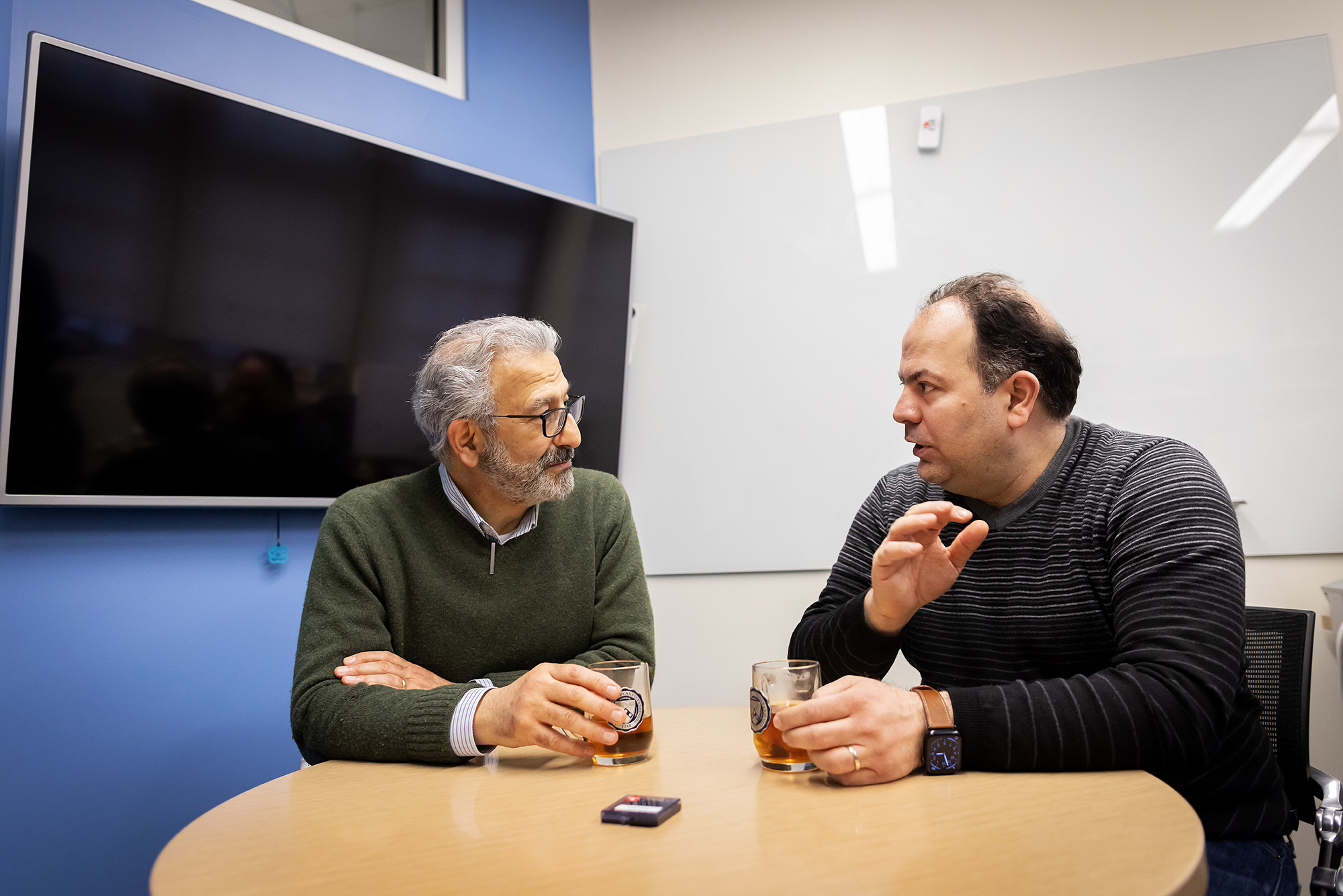 Nader Engheta and Firooz Aflatouni seated at a table drinking tea from Penn-branded mugs.