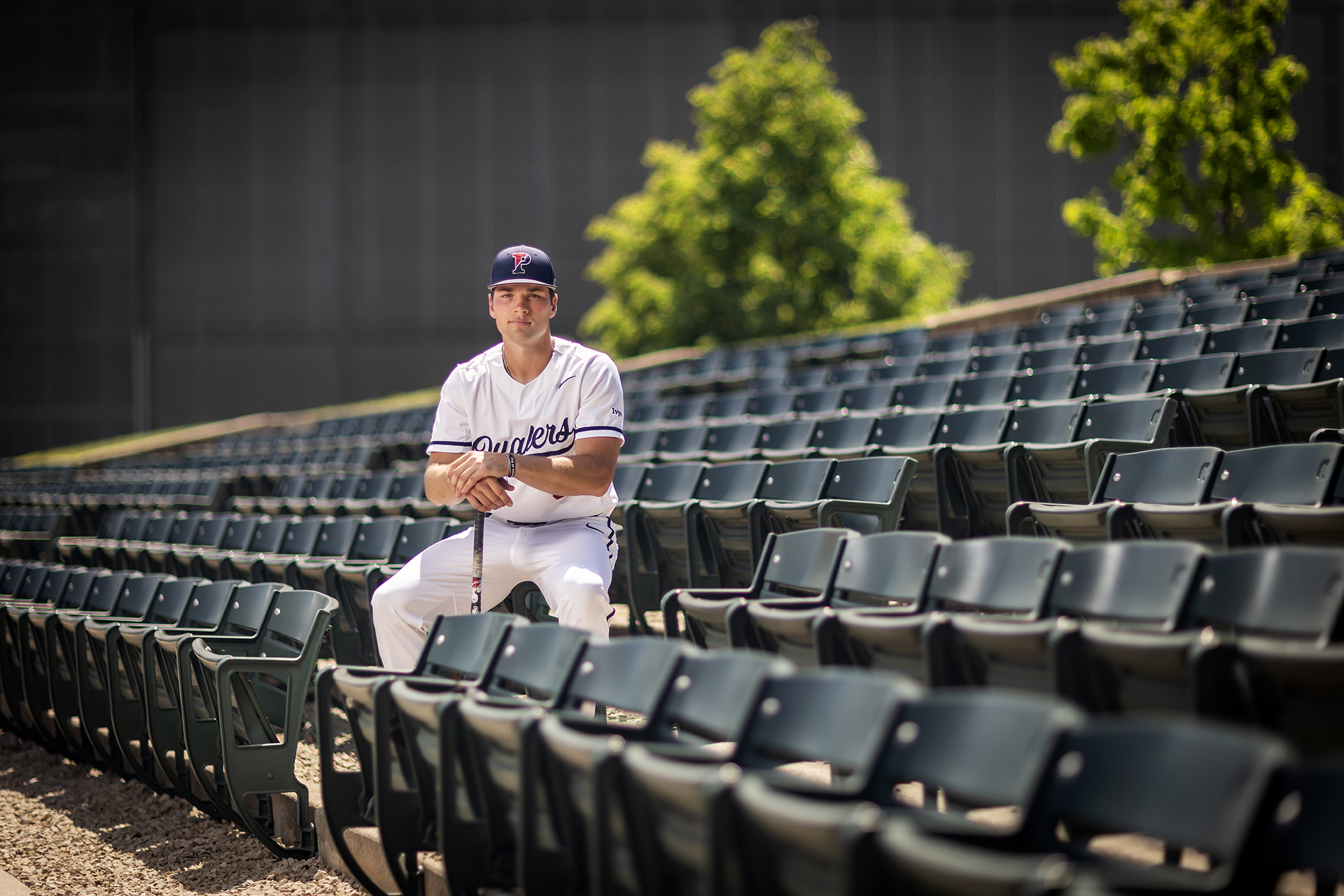 Wyatt Henseler sits in the bleachers at Meiklejohn Stadium while wearing his white uniform and leaning on a bat.