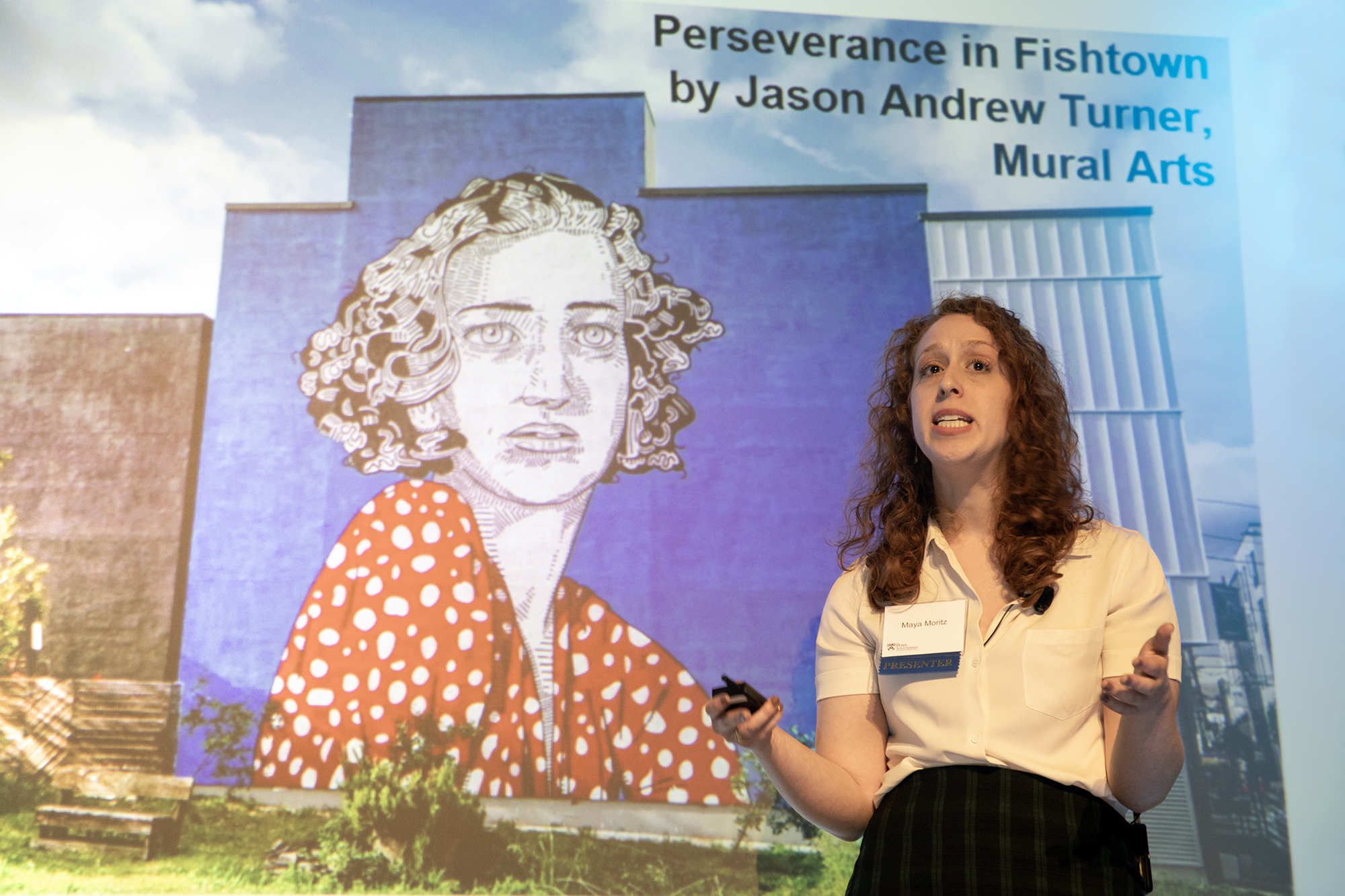 Maya Moritz giving a lecture in front of a mural.