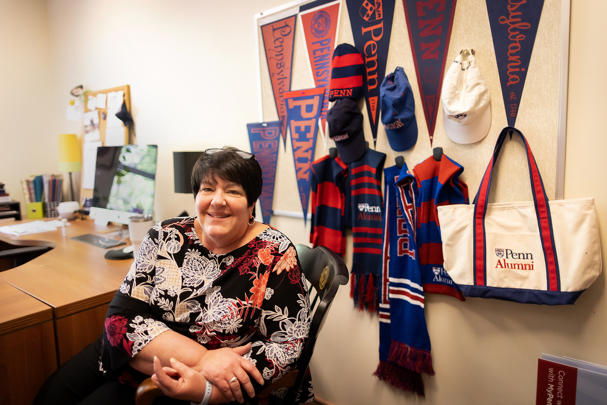 Kristina Clark sitting at a desk with Penn pennants, hats, flags, and bag behind her
