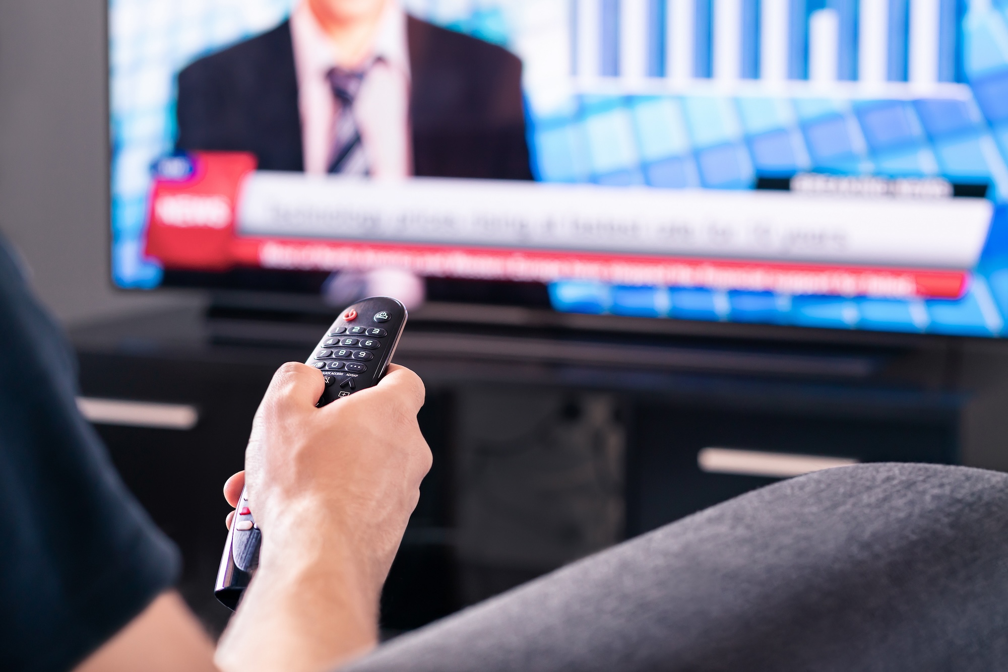 A person is sitting on a couch, holding a remote control in their right hand, and pointing it at a television displaying a news broadcast with a blurred image of an anchor and scrolling text at the bottom.