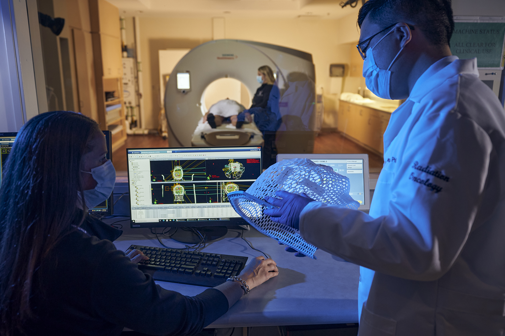 Two doctors looking at a mesh face shield in front of a computer with a person in a scan machine in background.