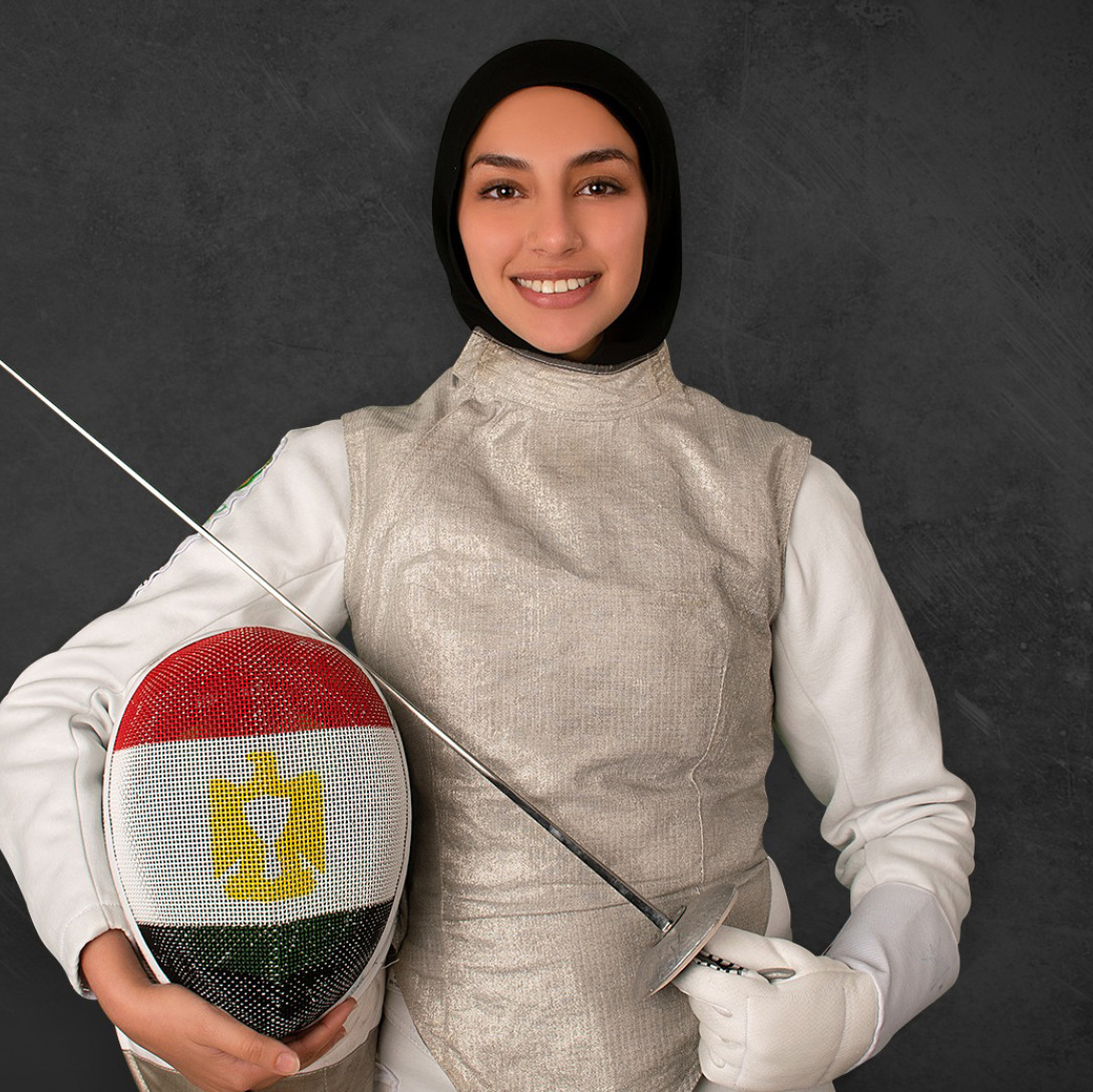 Malak Hamza stands in her fencing uniform holding her sword and helmet