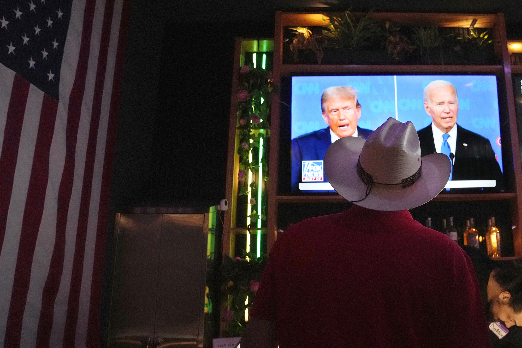 The back of a man wearing a white cowboy hat near an American flag can be seen in front of a television showing the first 2024 presidential debate between Biden and Trump.