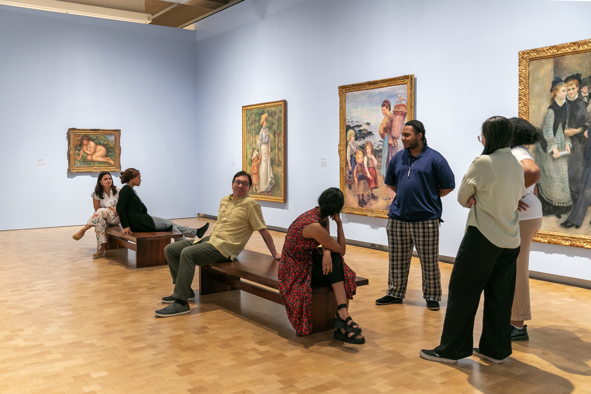 Museum interns stand and sit in a museum salon. The blue walls are hung with large Renoir paintings