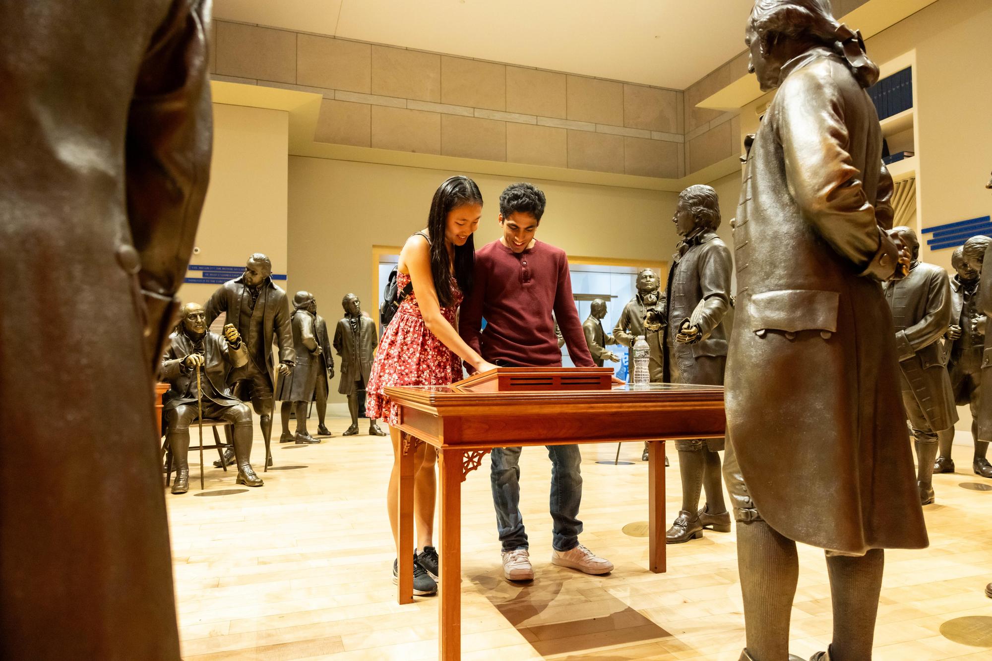 Two new Penn students in a museum room with many bronze founding fathers statues on NSO weekend.