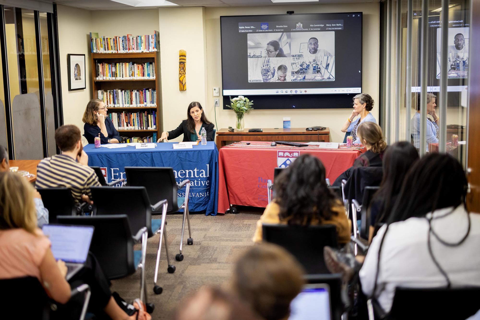 Three women sit at tables in front of an audience. A Zoom screen with three additional speakers is behind them.