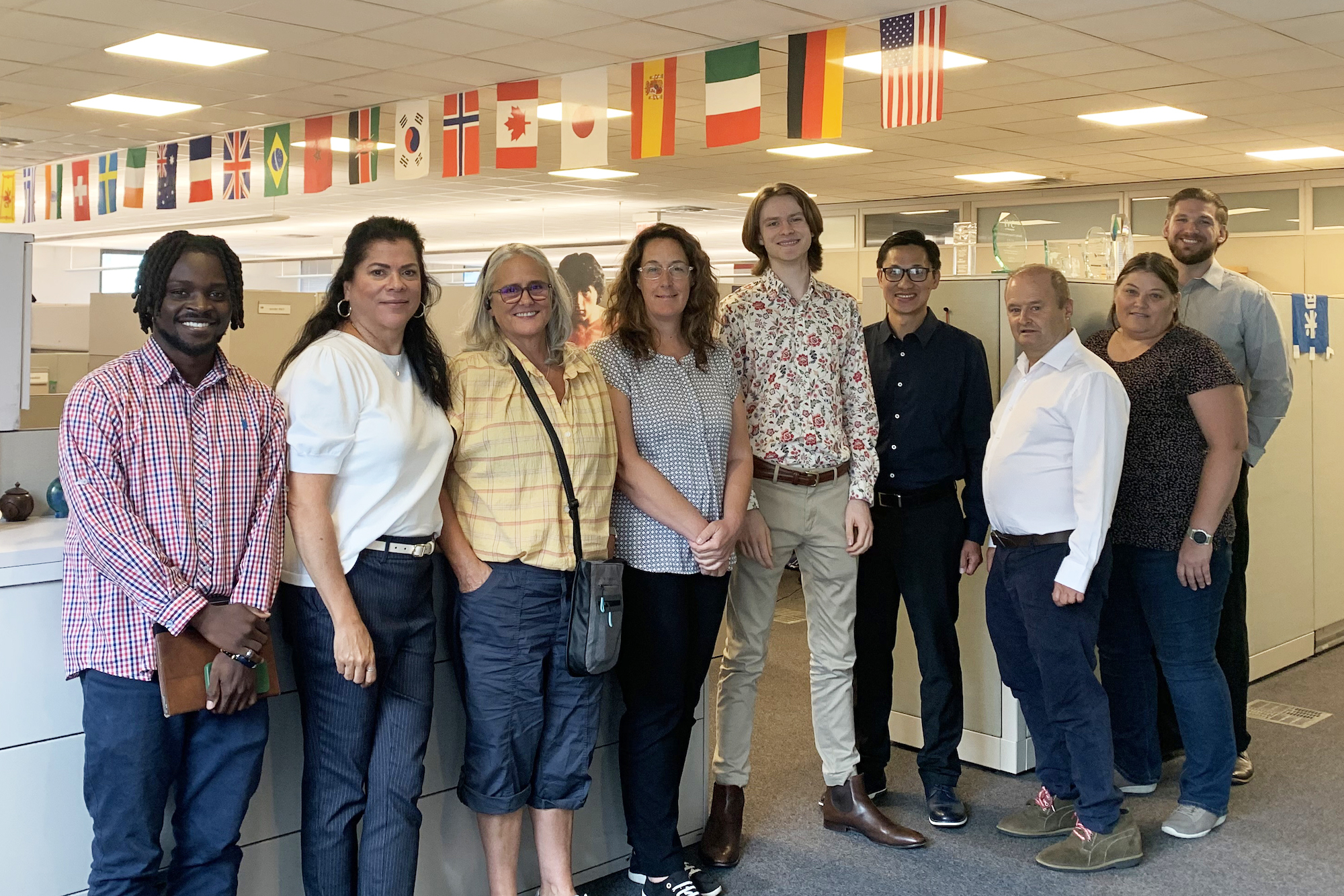 Nine people stand in front of office cubicles. Above them, a string of national flags