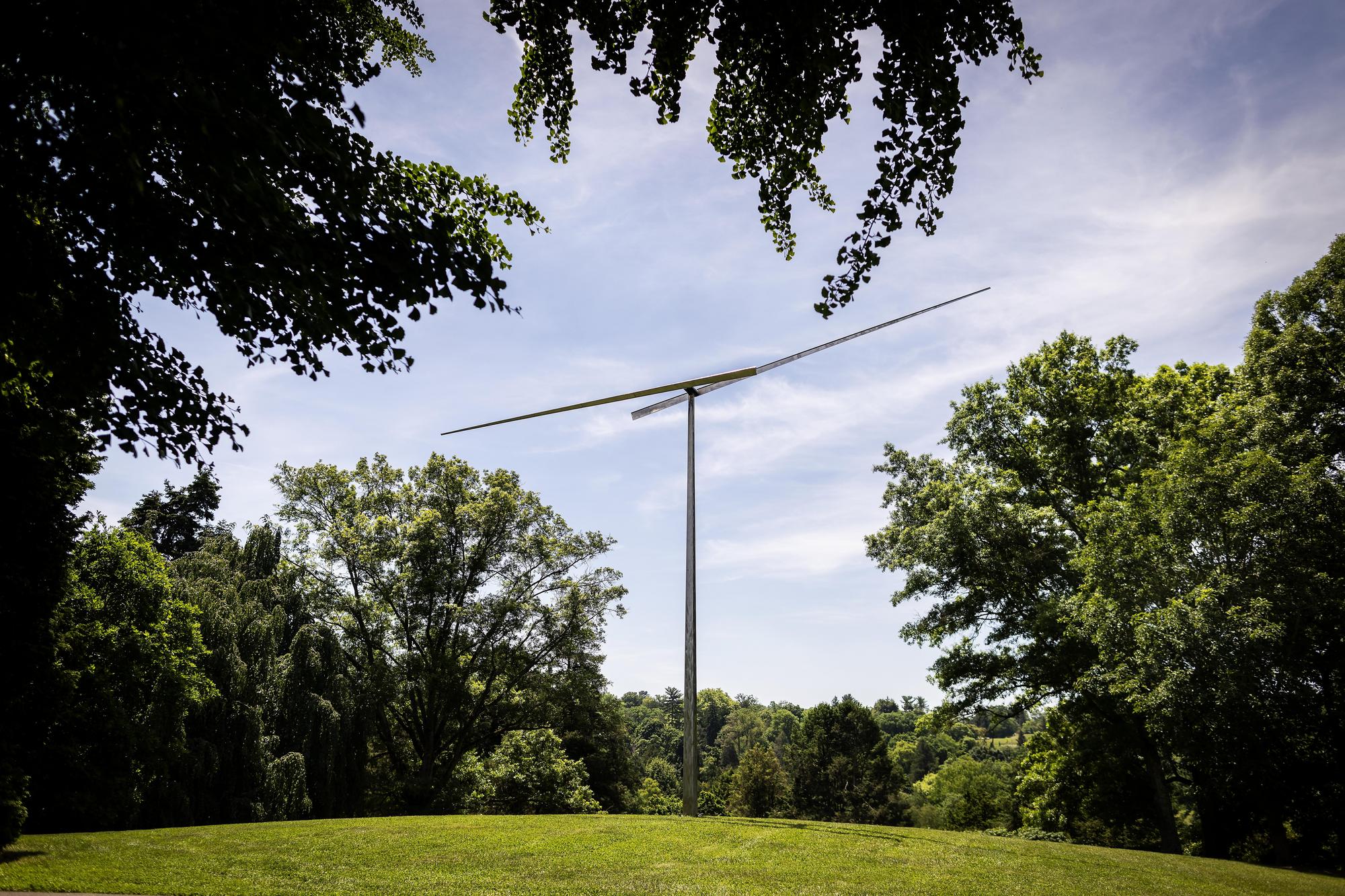 At the top of a sloping hill framed by trees, a grey metal sculpture looks like a watch face