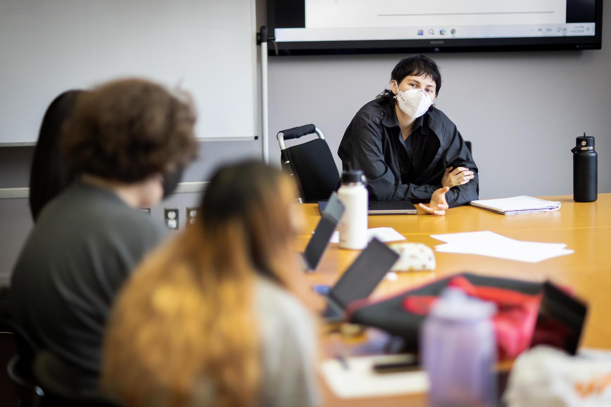 Mae Eskenazi, masked, sits at the head of a long table teaching students