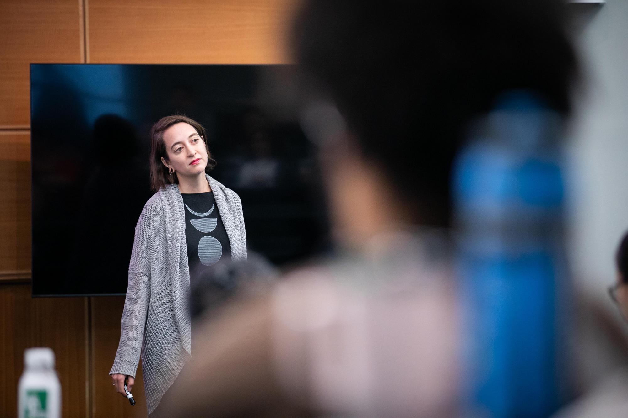 Jessa Lingel stands in front of a black screen, her head cocked to the side. In the foreground (blurred) are a student's head and water bottle.