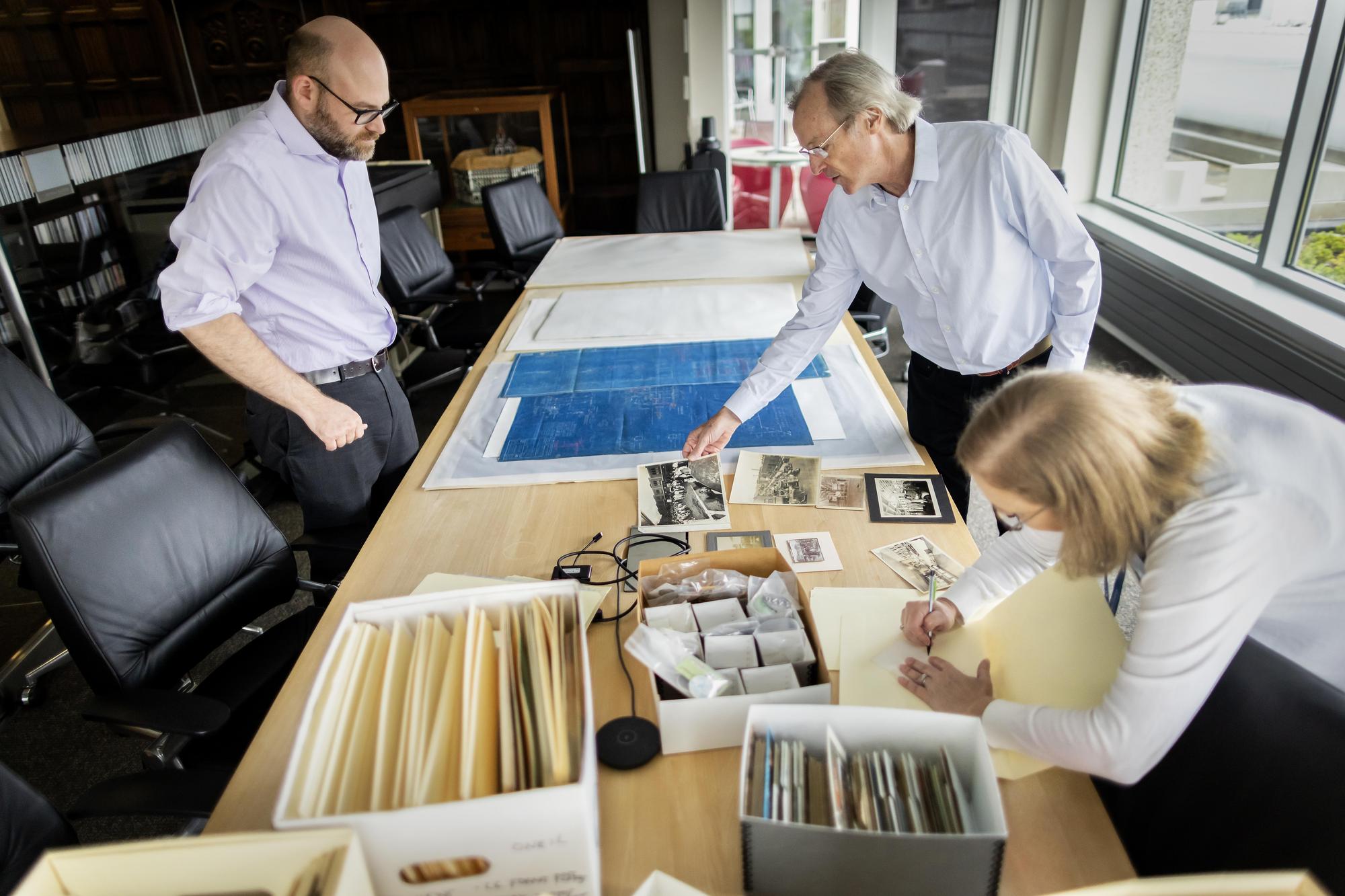 Three people looking at blueprints and postcards and other historic materials on a table in a room with windows.